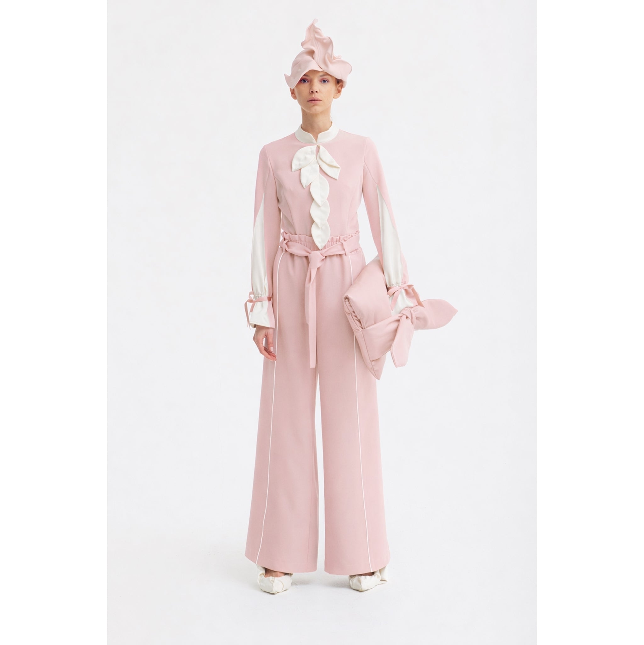 A person stands against a white background wearing JULIA ALLERT’s Elegant Fitted Blouse in pale pink, paired with wide-leg pants, a matching pink hat, and a large bow-accented clutch crafted from luxury suiting fabric.