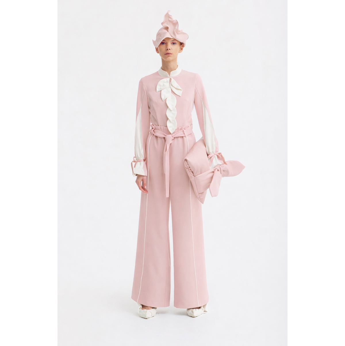 A person stands against a white background wearing JULIA ALLERT’s Elegant Fitted Blouse in pale pink, paired with wide-leg pants, a matching pink hat, and a large bow-accented clutch crafted from luxury suiting fabric.