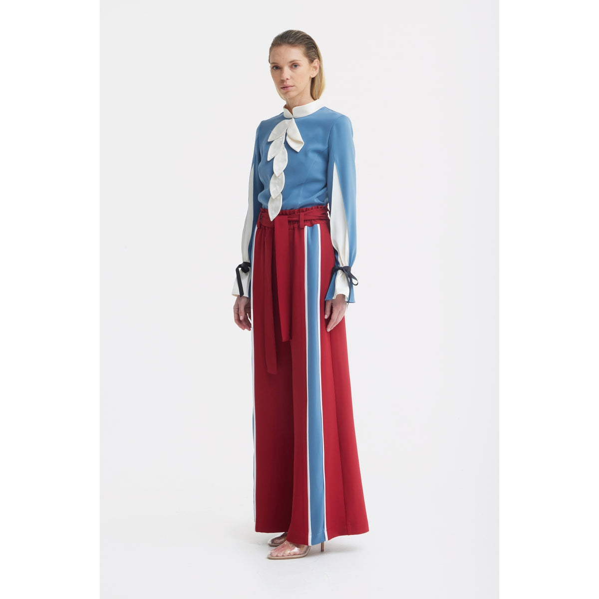 A woman with light brown hair stands against a white background wearing the JULIA ALLERT Elegant Fitted Blouse - Blue with white accents and wide-leg red pants featuring blue and white stripes, paired with clear open-toe shoes.