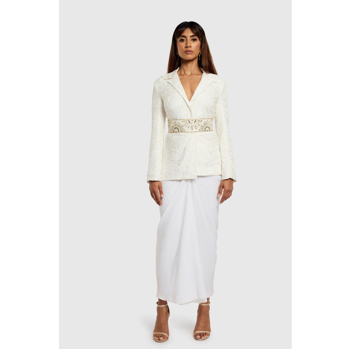 A woman faces forward in a modern luxury look, wearing INTERNATIONAL APPEAL’s White Paisley Textured Blazer (PR Sample) paired with a long white skirt and heeled sandals. Her long dark hair contrasts against the plain light background.