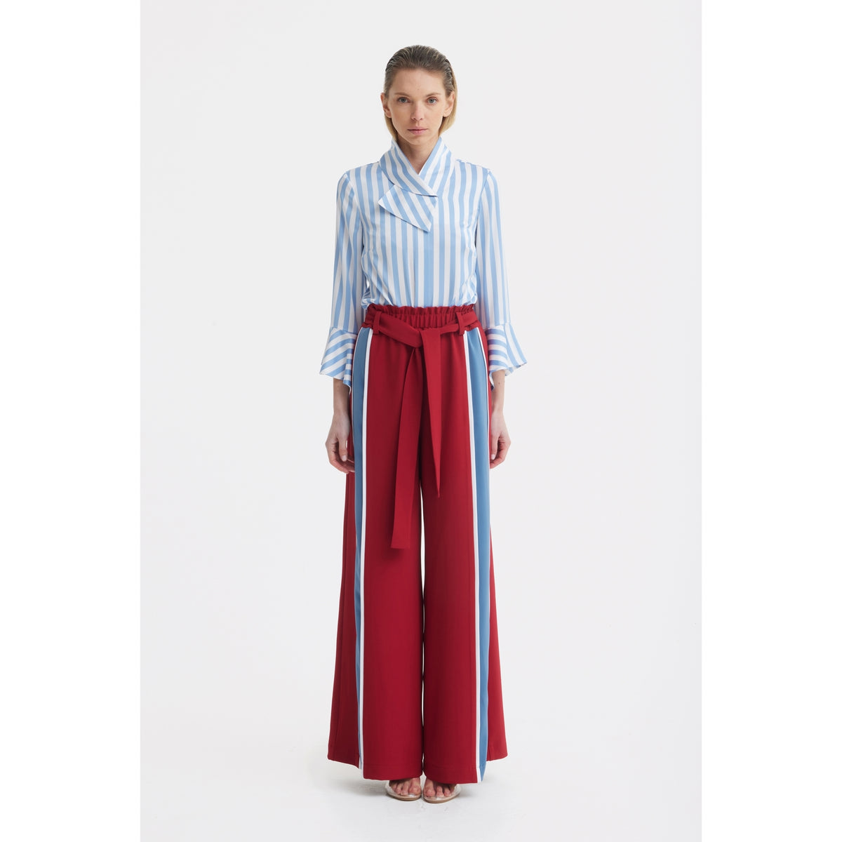 A woman faces forward in a JULIA ALLERT Satin Striped Blouse With Asymmetric Collar in light blue and white, paired with wide red high-waisted pants featuring a matching belt and light blue vertical stripes, against a plain white background.