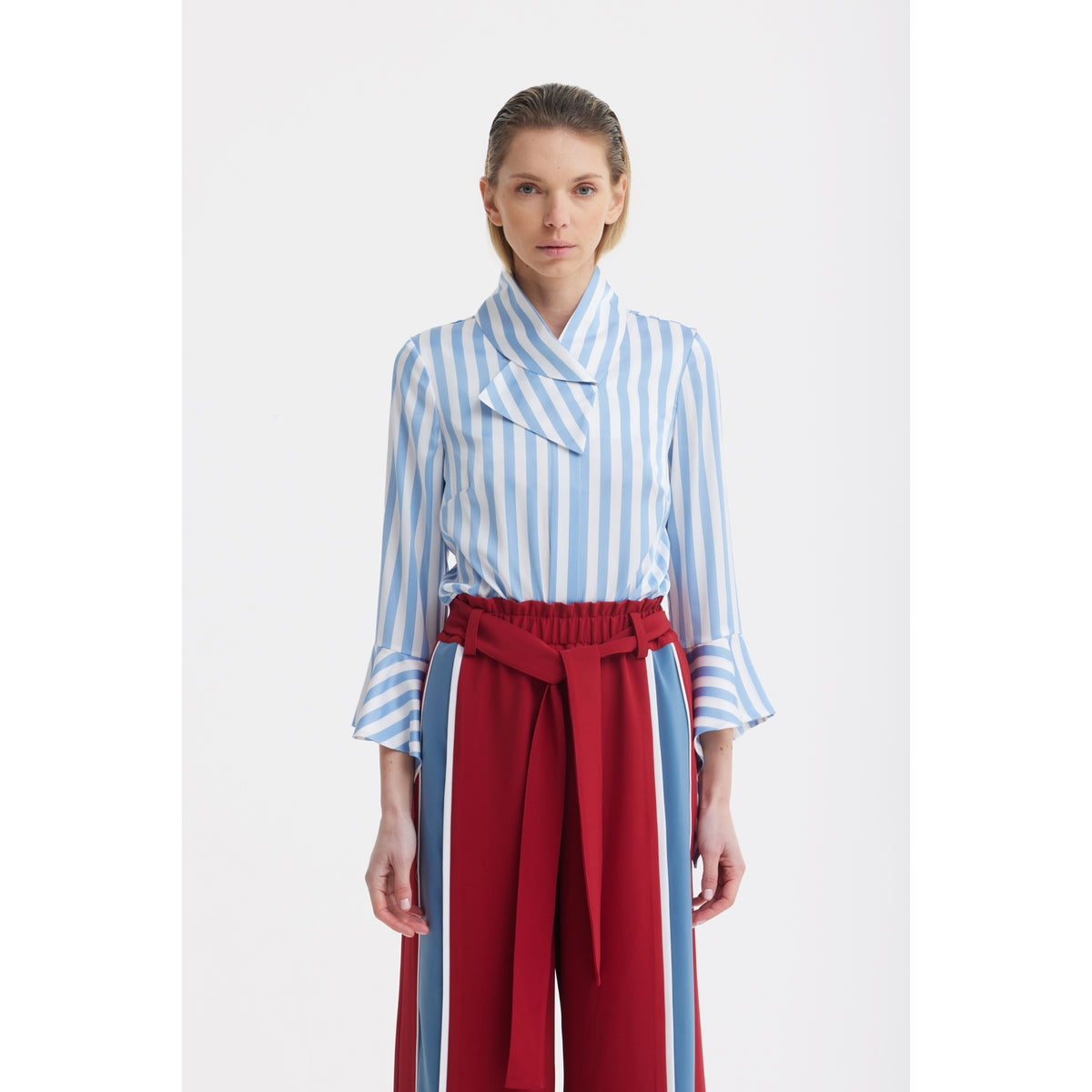 A woman faces forward in a JULIA ALLERT Satin Striped Blouse With Asymmetric Collar in light blue, paired with high-waisted red paperbag trousers featuring a matching belt.