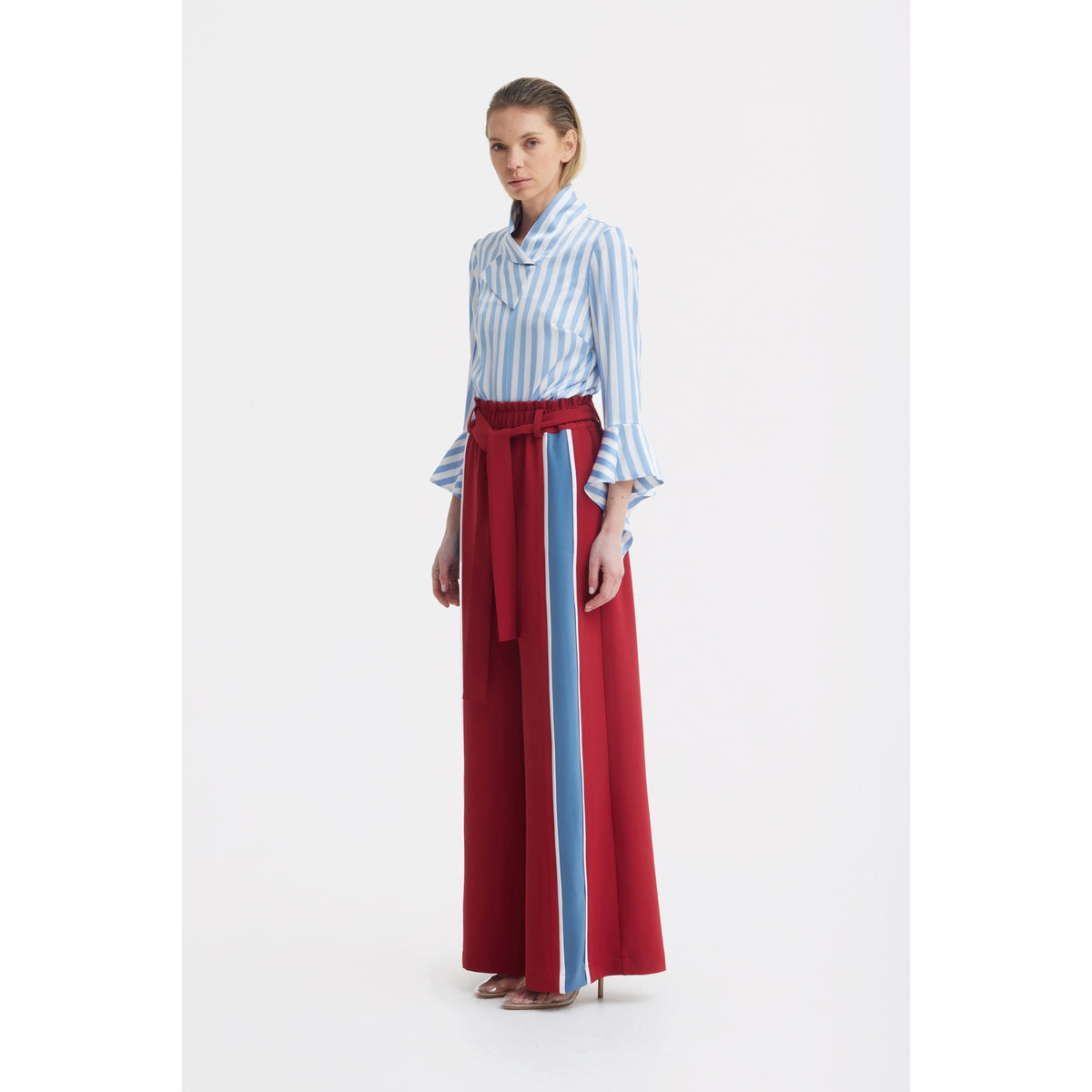 A woman stands wearing a JULIA ALLERT Satin Striped Blouse With Asymmetric Collar in light blue, paired with high-waisted red wide-leg pants with a vertical blue stripe, a matching red belt, and high-heeled shoes.