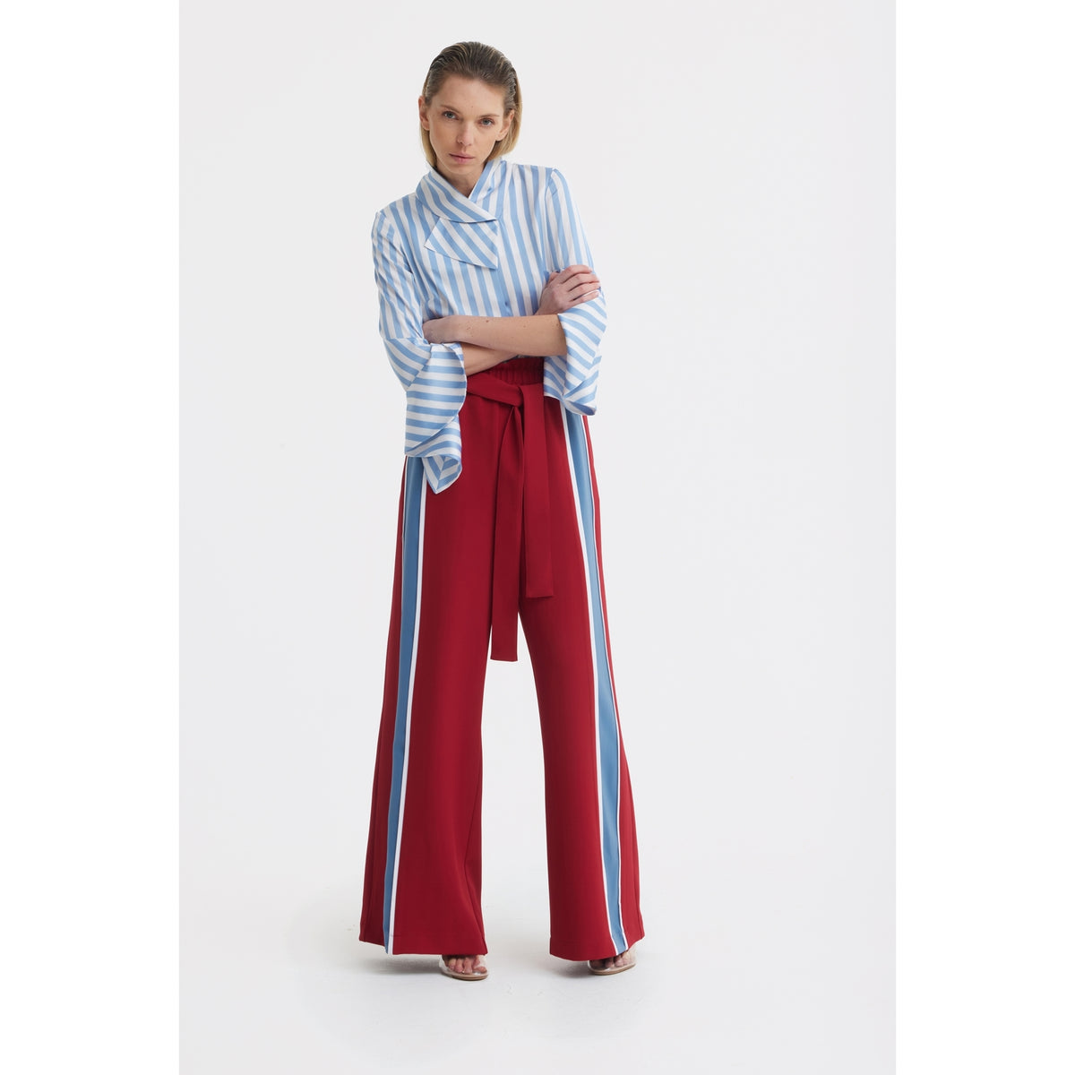 A woman stands with arms crossed wearing a JULIA ALLERT Satin Striped Blouse With Asymmetric Collar in light blue and high-waisted red wide-leg pants with blue and white side stripes, set against a plain white background.