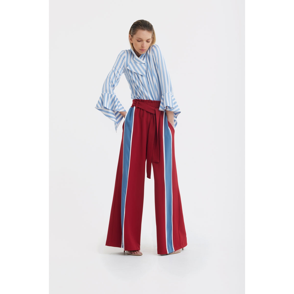 A woman poses in a JULIA ALLERT Satin Striped Blouse With Asymmetric Collar in light blue, tucked into high-waisted, wide-leg red pants with vertical blue and white stripes and a matching belt.