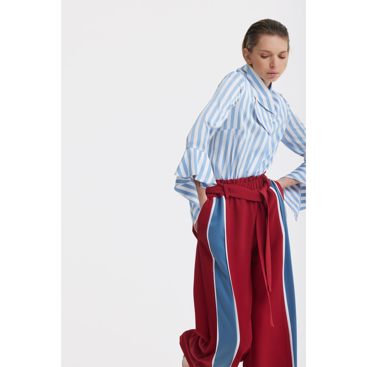 A person wearing the JULIA ALLERT Satin Striped Blouse With Asymmetric Collar in light blue and high-waisted red pants with blue and white side stripes stands against a white background, looking down with a neutral expression.