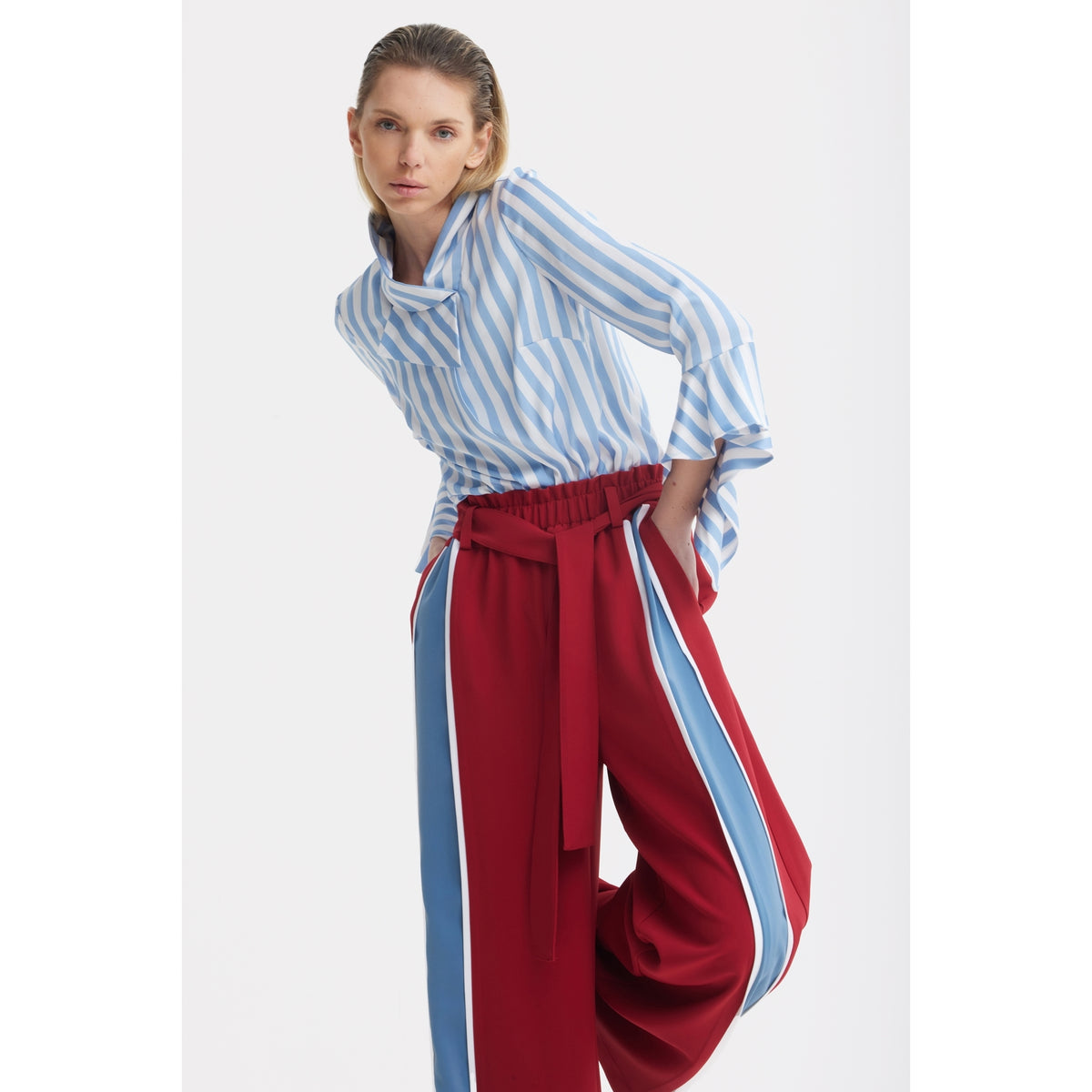 A person wears the JULIA ALLERT Satin Striped Blouse With Asymmetric Collar in light blue, paired with loose red pants with wide blue and white side stripes, posing against a plain white background.