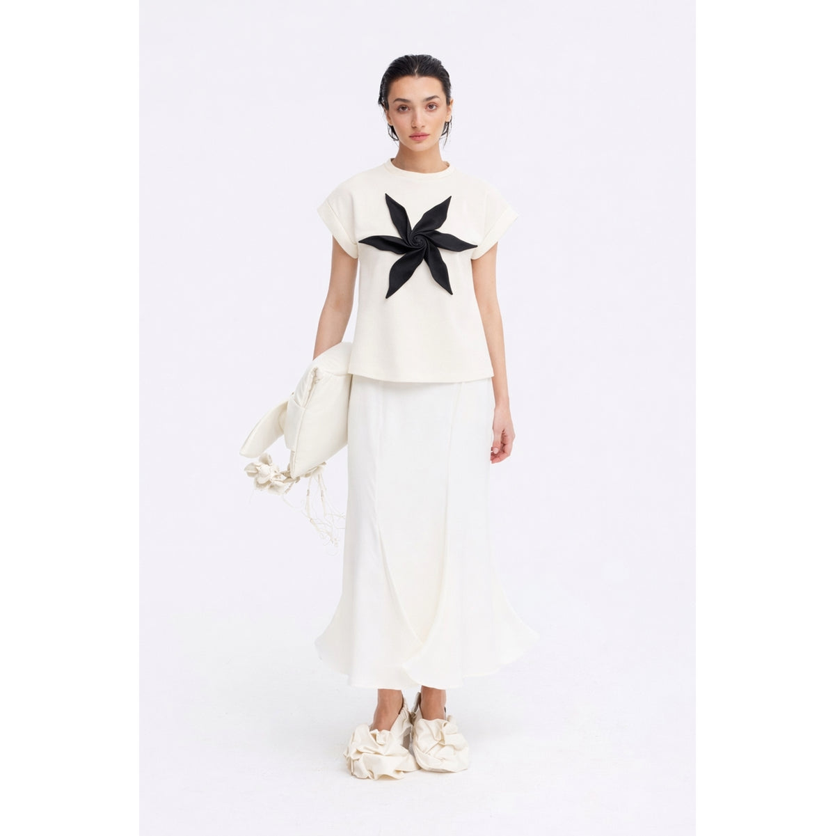 A woman stands against a plain white background wearing the JULIA ALLERT Crew-Neck Jersey T-Shirt With Flower in Black, paired with a long white skirt, white ruffled shoes, and holding a textured white clutch bag.
