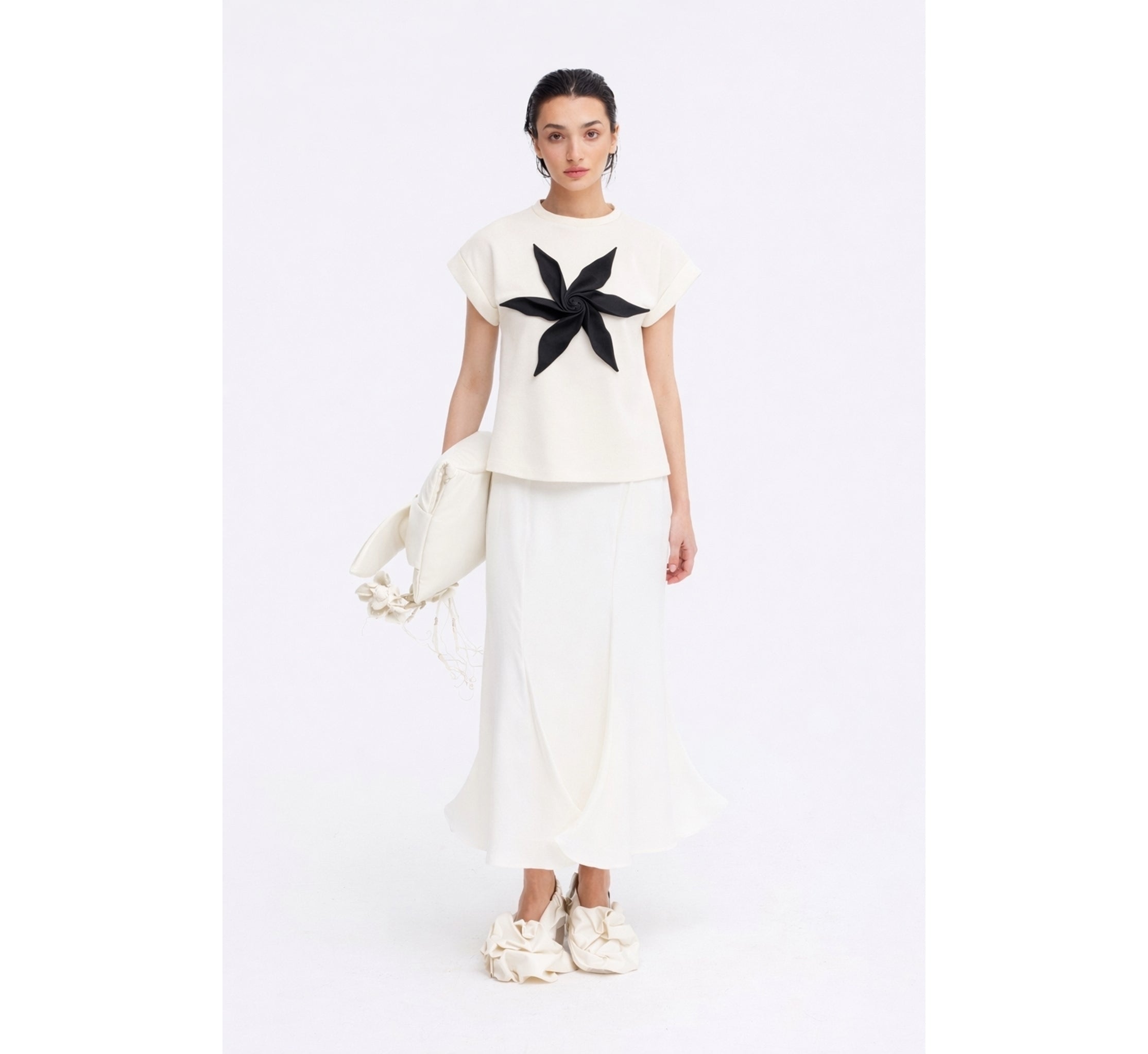 A woman poses against a white background in a JULIA ALLERT white top with a bold black flower, the High Waist Double-Sided Satin Maxi Skirt – Ivory, ruffled white shoes, and a white bag with decorative balls—an eveningwear standout.