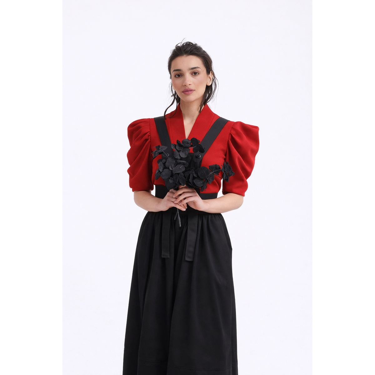 A woman in a JULIA ALLERT V-Neck Puff Sleeve Top - Red and a black skirt holds a bouquet of black flowers against a white background.
