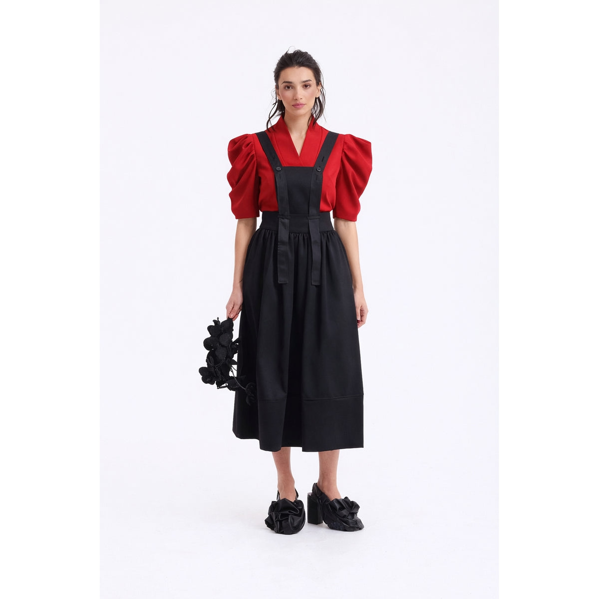 A woman stands against a white background wearing the JULIA ALLERT V-Neck Puff Sleeve Top in red under a black pinafore-style dress, accessorized with a black floral piece and matching black ruffled shoes.
