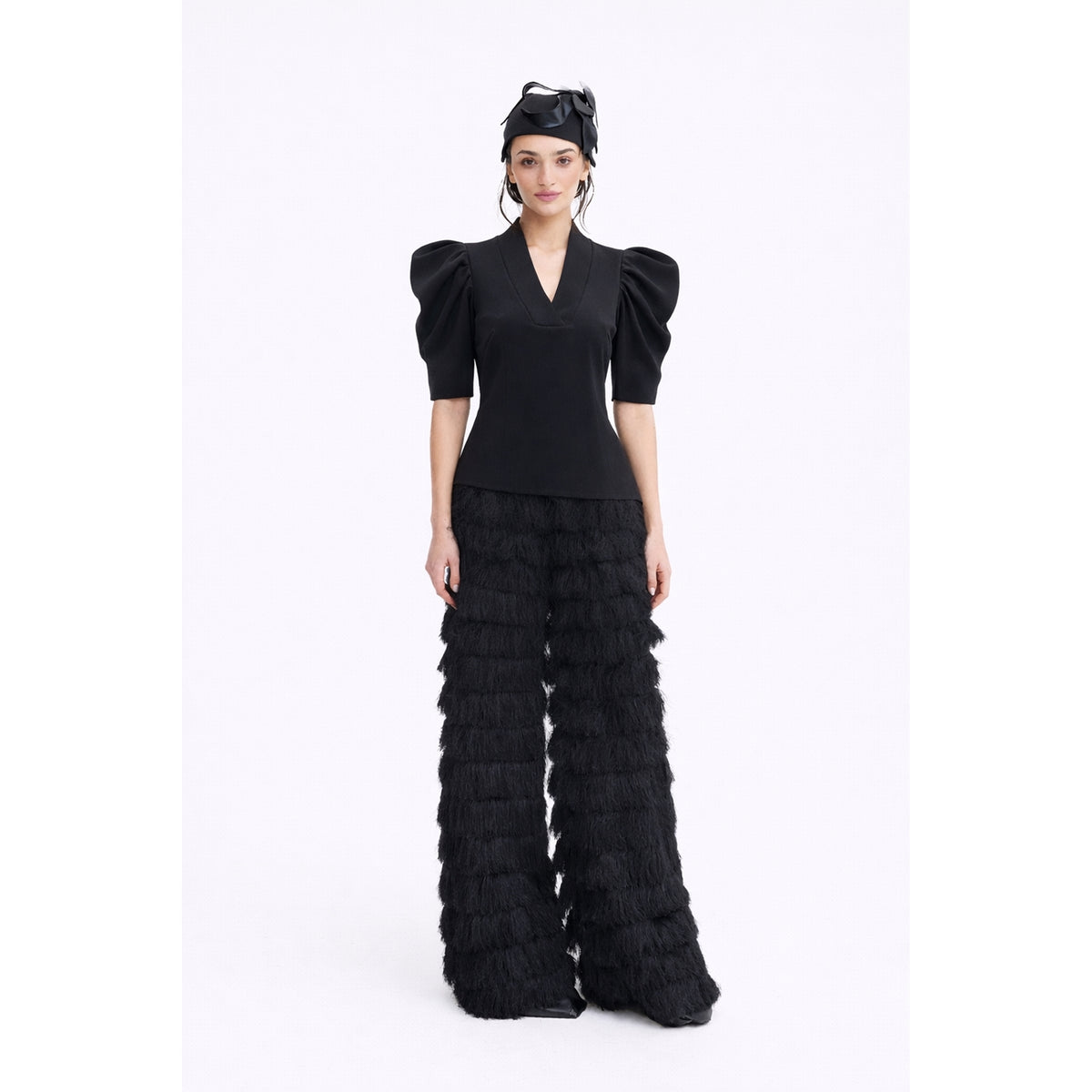 A woman stands against a white background wearing the JULIA ALLERT V-Neck Puff Sleeve Top in black, paired with tiered, fringed black wide-leg pants and a black bow headpiece.