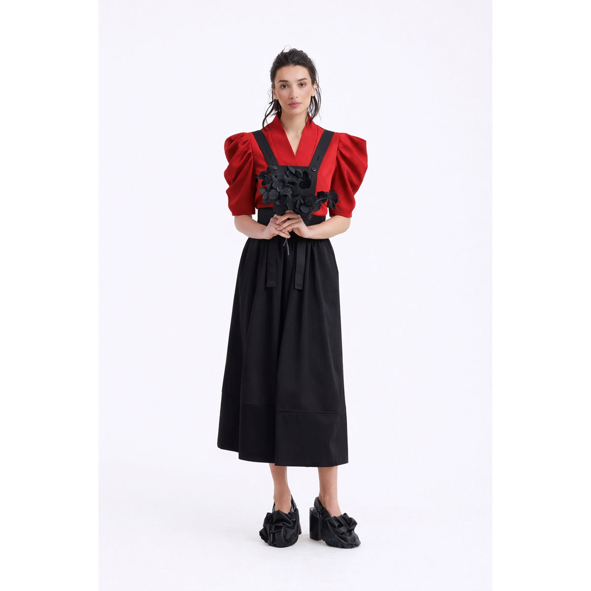 A woman stands against a white background wearing the JULIA ALLERT V-Neck Puff Sleeve Top - Red, paired with a black wrap-style dress featuring floral accents at the waist and black heeled shoes adorned with fabric details.
