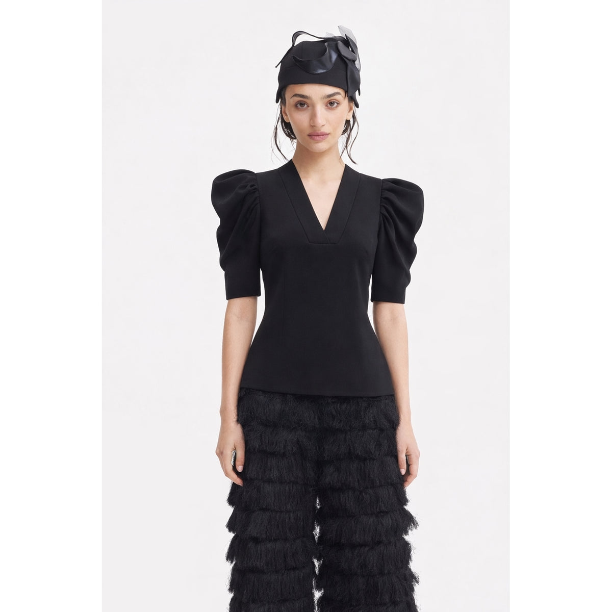 A woman stands against a white background wearing the JULIA ALLERT V-Neck Puff Sleeve Top in black, paired with black tiered fringe pants and a black decorative hat.