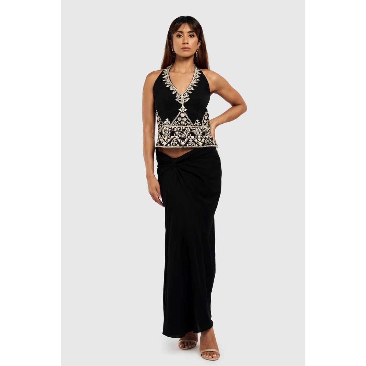 Against a light gray background, a woman wears the INTERNATIONAL APPEAL Black Floral-Embroidered Tailored Vest (PR Sample) styled with a long black knotted skirt and open-toe heels, exuding contemporary luxury.