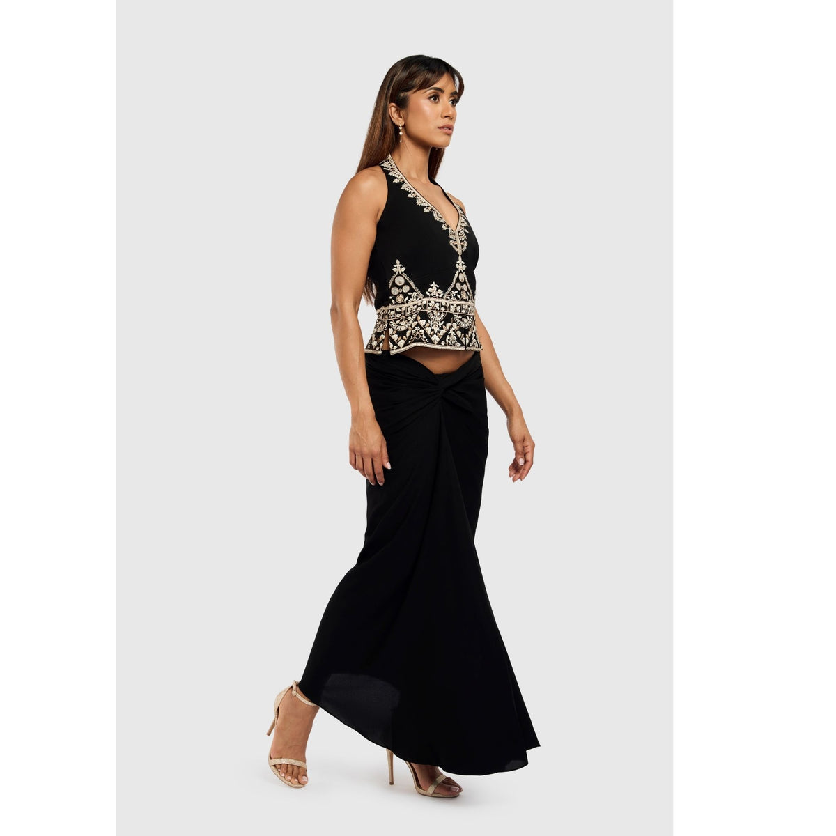 A woman with long brown hair wears the INTERNATIONAL APPEAL Black Floral-Embroidered Tailored Vest (PR Sample), a long black skirt, gold earrings, and beige high-heeled sandals as she walks in profile against a light background.