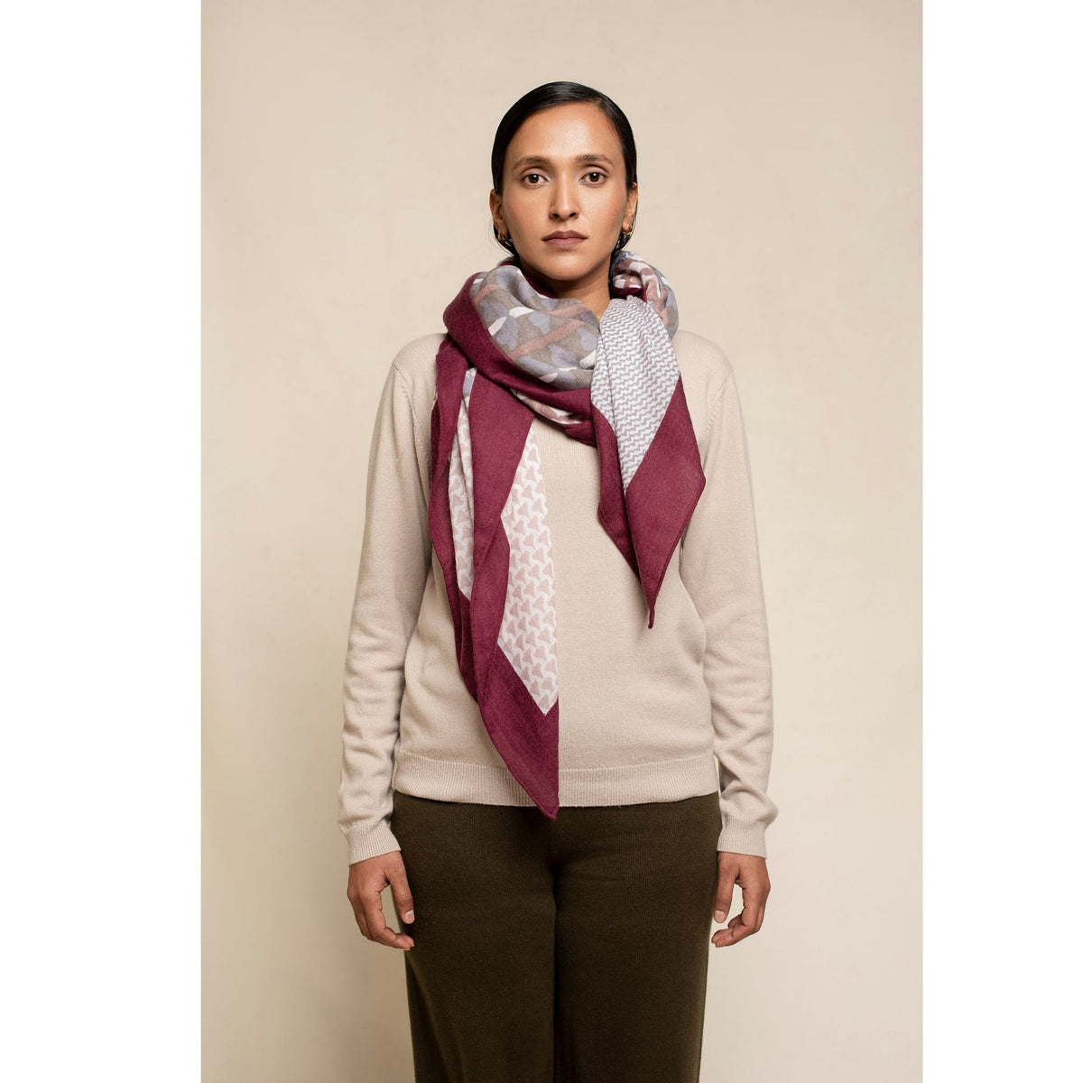 A woman with dark hair tied back wears a beige sweater, olive pants, and the OO-TO Printed Square Scarf Micro Pattern Patch with Red Border. She stands facing forward against a plain light background, embodying sustainable luxury.