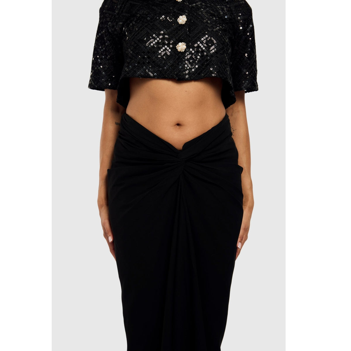 A person wears a black sequined crop top and the Black Sari-Draped Maxi Skirt by INTERNATIONAL APPEAL, featuring georgette draping and a waist knot for a modern sari look. The midriff is exposed, set against a plain light gray background.