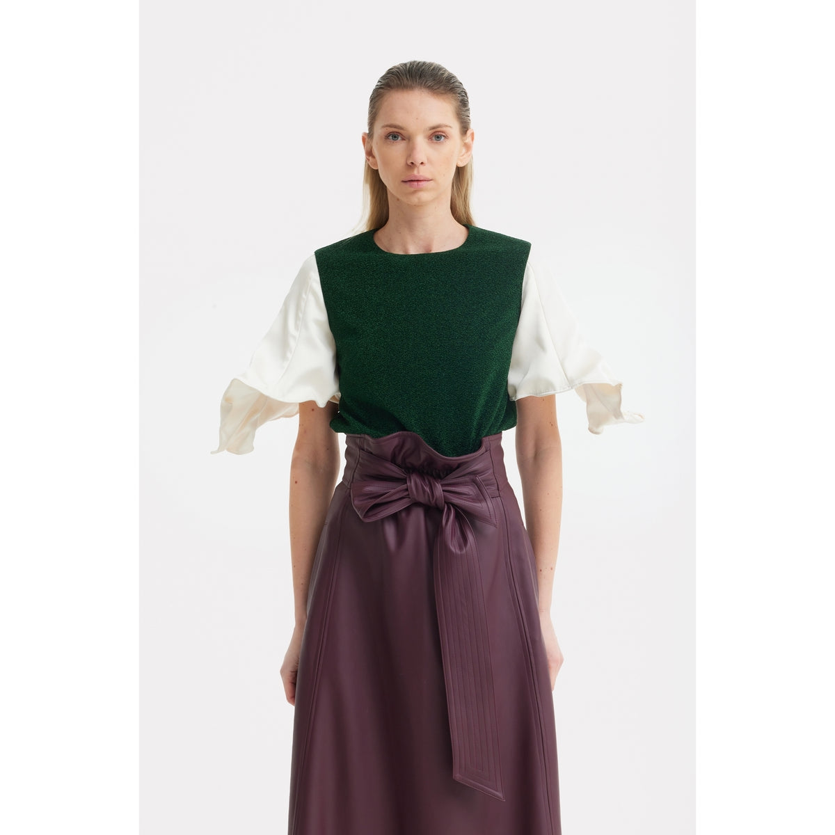 A woman faces forward in a JULIA ALLERT Green Lurex Knit Top With Adjustable Satin Sleeves, layered over a white blouse and paired with a high-waisted plum skirt with a large bow. She has straight, light brown hair.