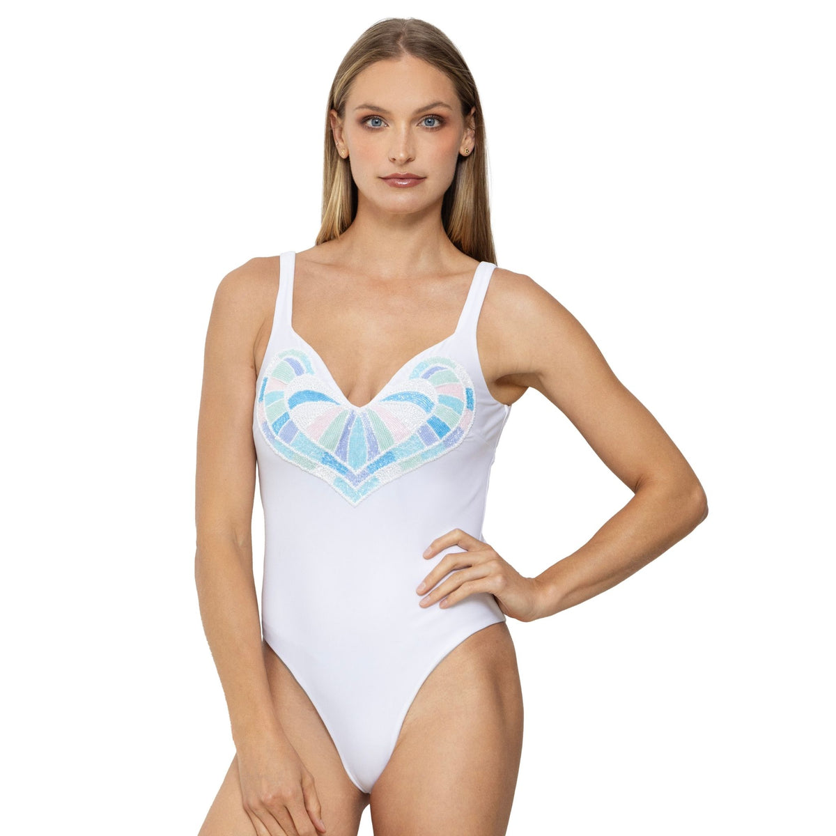 A woman with long blonde hair models the SELF-ADORE Amor Hand-Beaded Embellished One-Piece Swimsuit in White, showcasing its colorful geometric heart detail, standing with one hand on her hip against a white background.