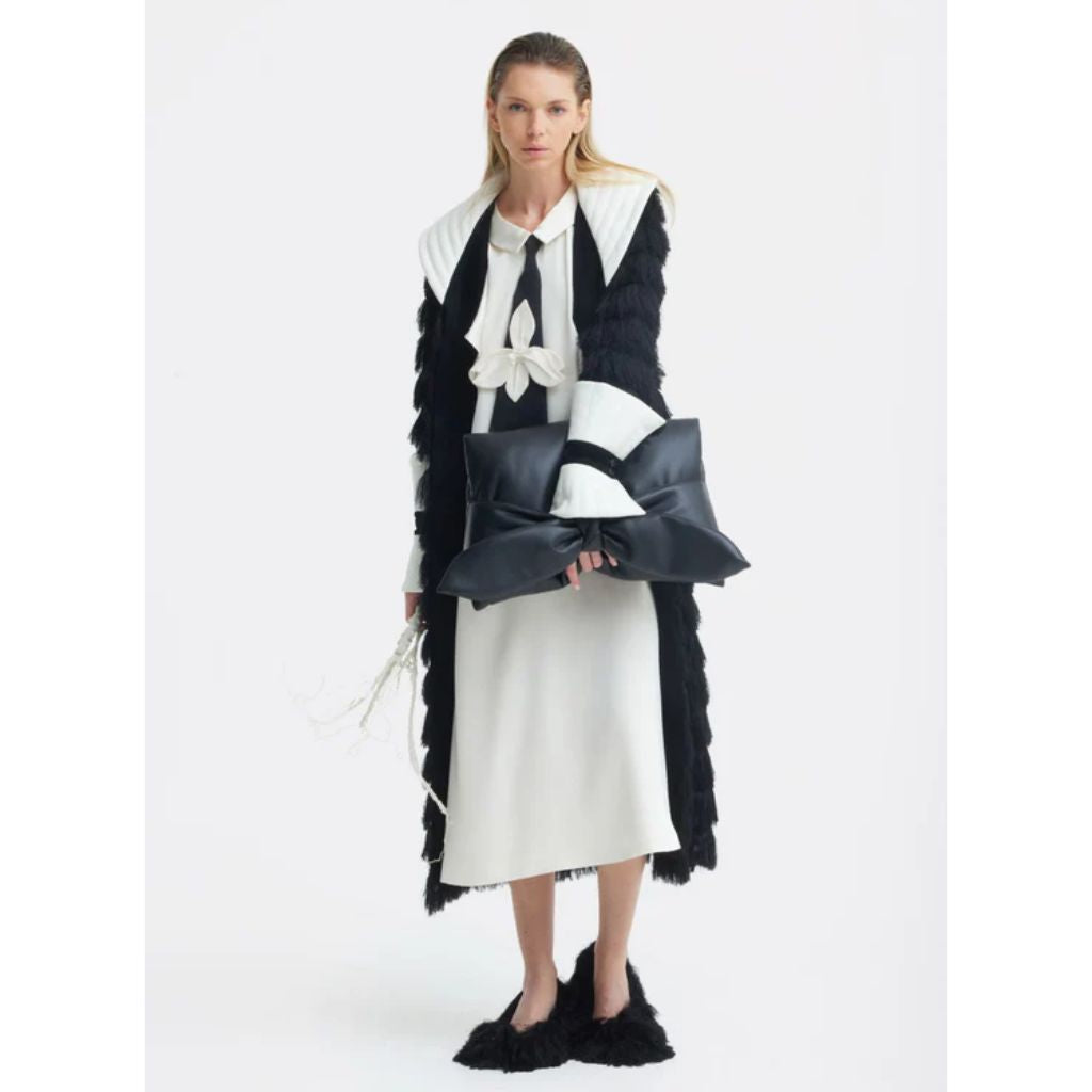 A woman in a JULIA ALLERT Double-Layer Satin Midi Dress With Tie Detail – Ivory stands against a plain background, pairing the premium satin dress with an oversized black bow clutch, furry black shoes, and a textured black coat.