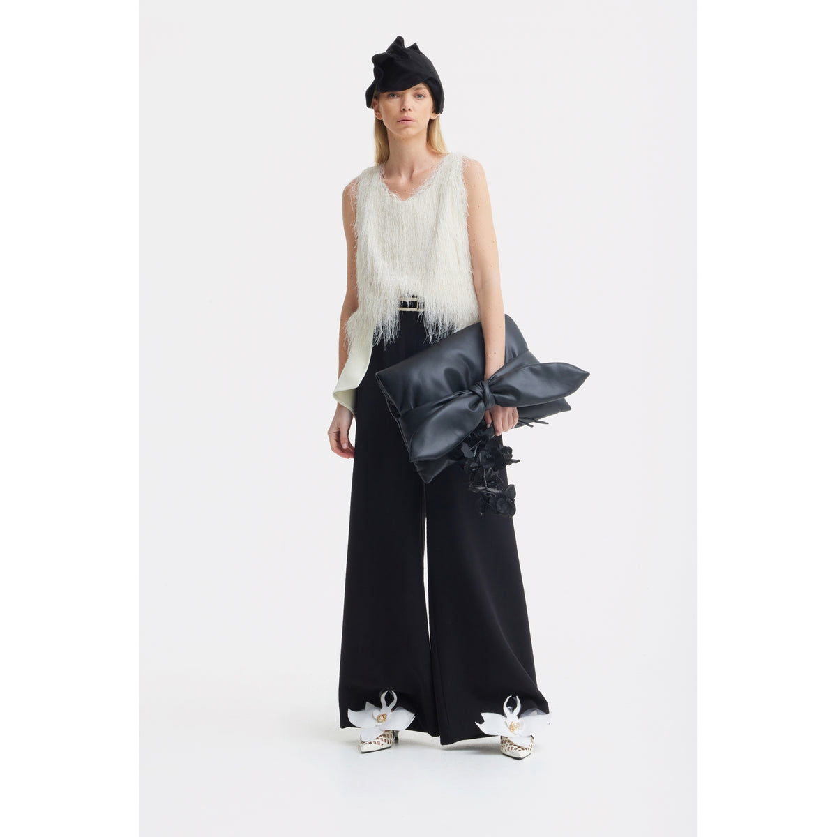 A woman in a black beret, sleeveless white fringe top, and JULIA ALLERT Statement Waist Wide-Leg Trousers in black holds a large black bow-shaped clutch and finishes her look with embellished white shoes.