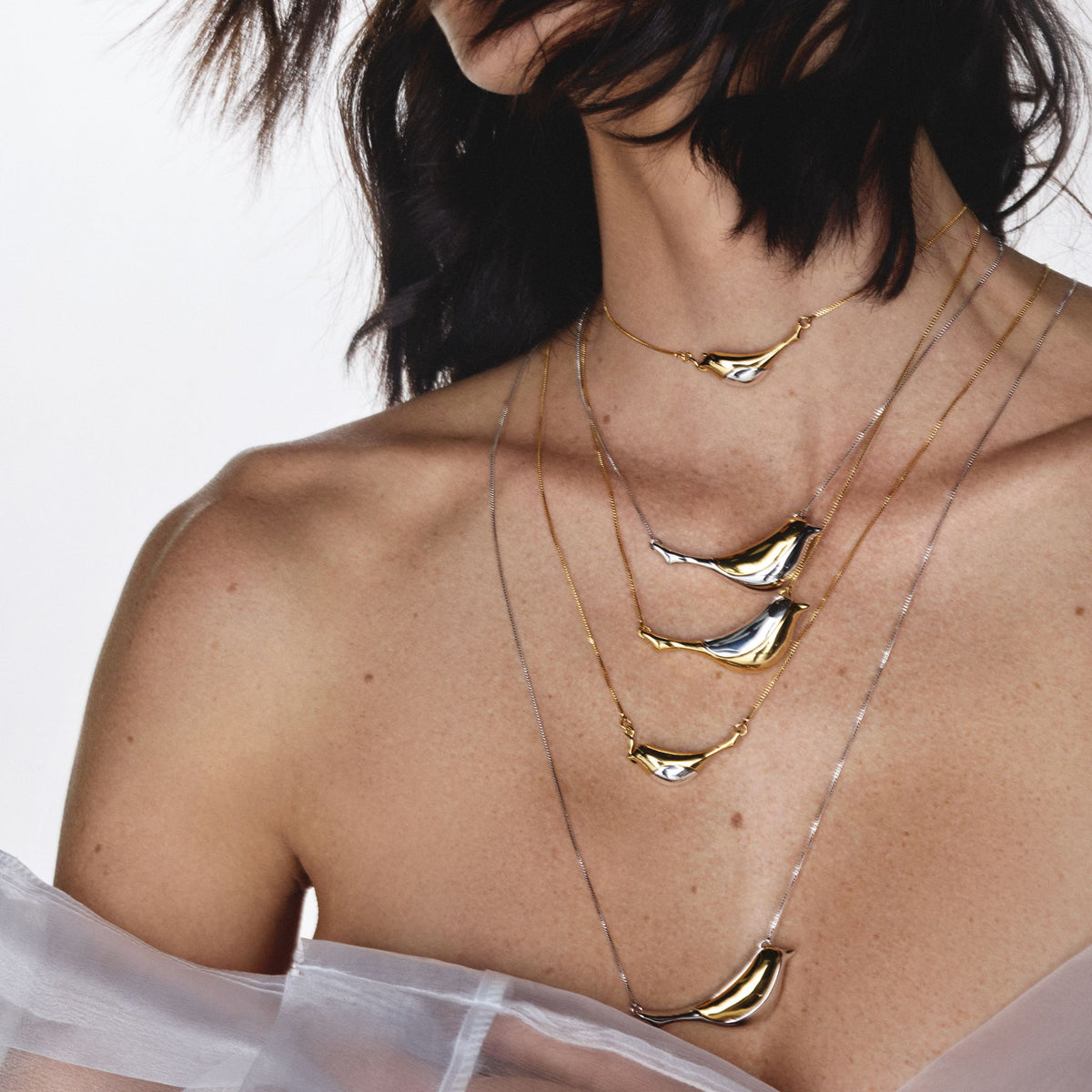 A woman wears four layered necklaces, including the ANNA PRATA Silver Aviva Necklace from the Aviva Collection, featuring curved gold and silver pendants. Her dark hair and bare shoulders are draped in sheer white fabric against a white background.