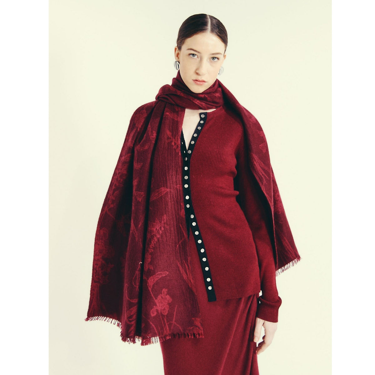 A woman with tied-back hair wears an OO-TO Modern Flower Printed Woven Cashmere Scarf in deep red over a matching ribbed cardigan with black and white buttons and a red skirt, standing against a light background.