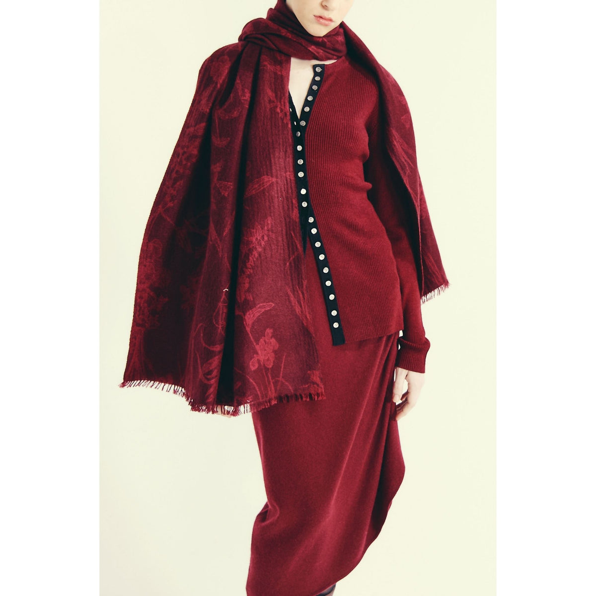 A woman in a deep red cardigan and skirt stands against a plain background, draped in OO-TO’s Modern Flower Printed Woven Cashmere Scarf—a statement of sustainable luxury. Her face is partially out of frame.