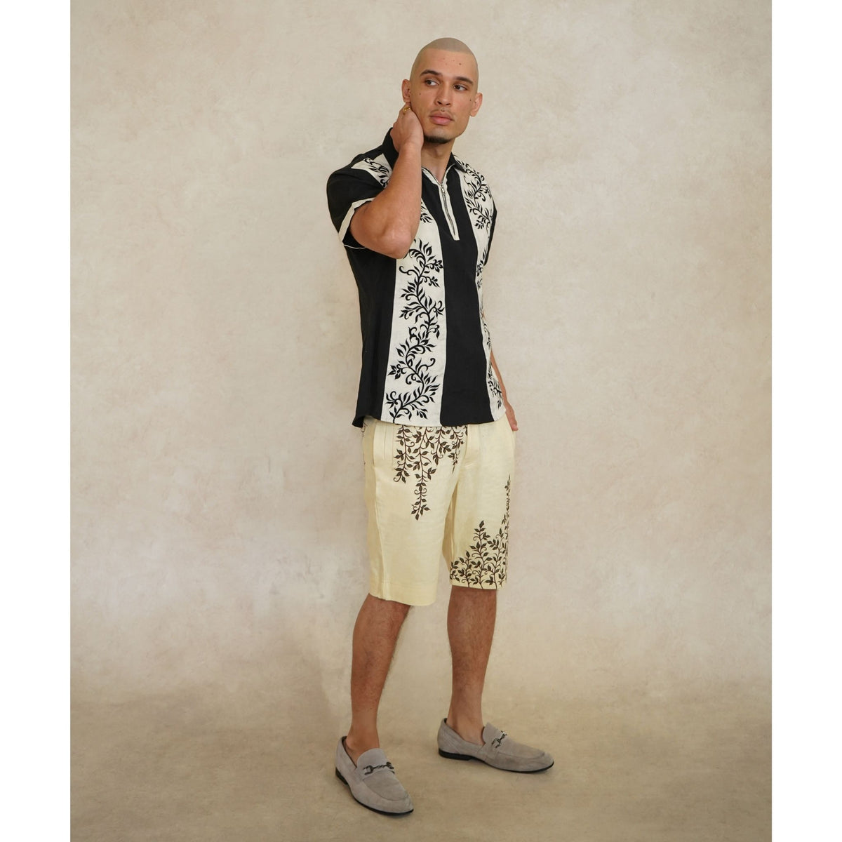 A man with a shaved head poses against a beige background, wearing INTERNATIONAL APPEAL's Black Floral-Embroidered Panel Shirt (PR Sample), paired with cream shorts and gray loafers. He stands with one hand touching his neck.