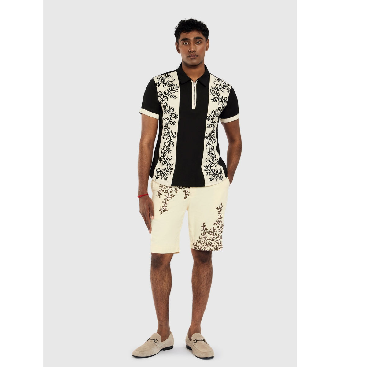 A man faces forward wearing INTERNATIONAL APPEAL’s Black Floral-Embroidered Panel Shirt (PR Sample) paired with cream shorts featuring matching floral designs. He stands with one hand in his pocket and beige loafers.