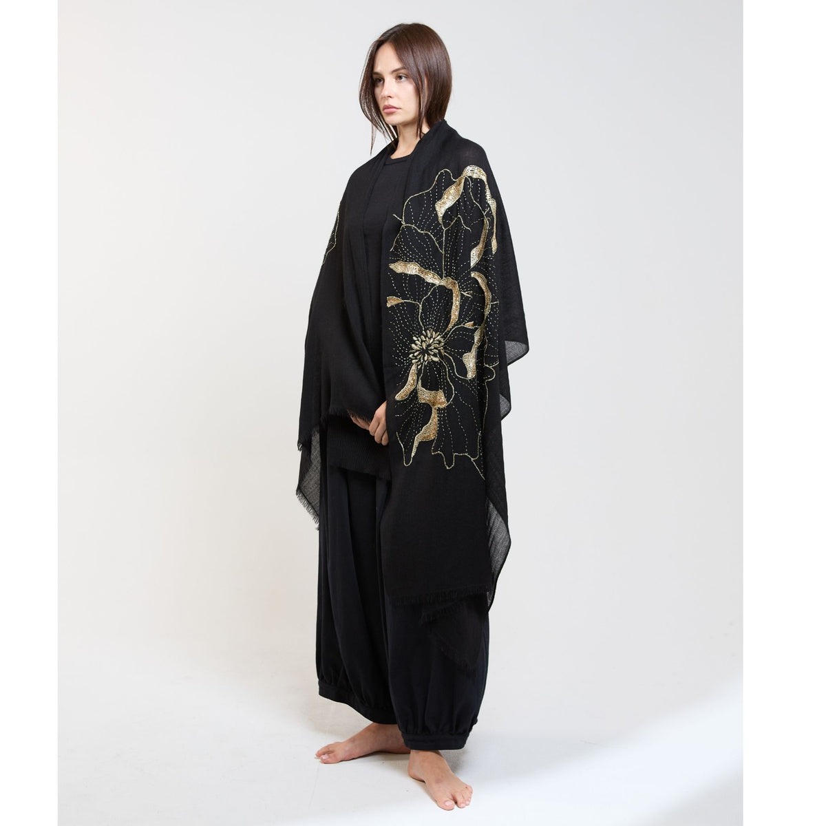 A woman with straight brown hair stands barefoot in loose black clothing and an OO-TO Black Blythe Flower Embroidered Stole—a luxurious handmade Nepalese scarf with gold floral embroidery—against a plain white background.