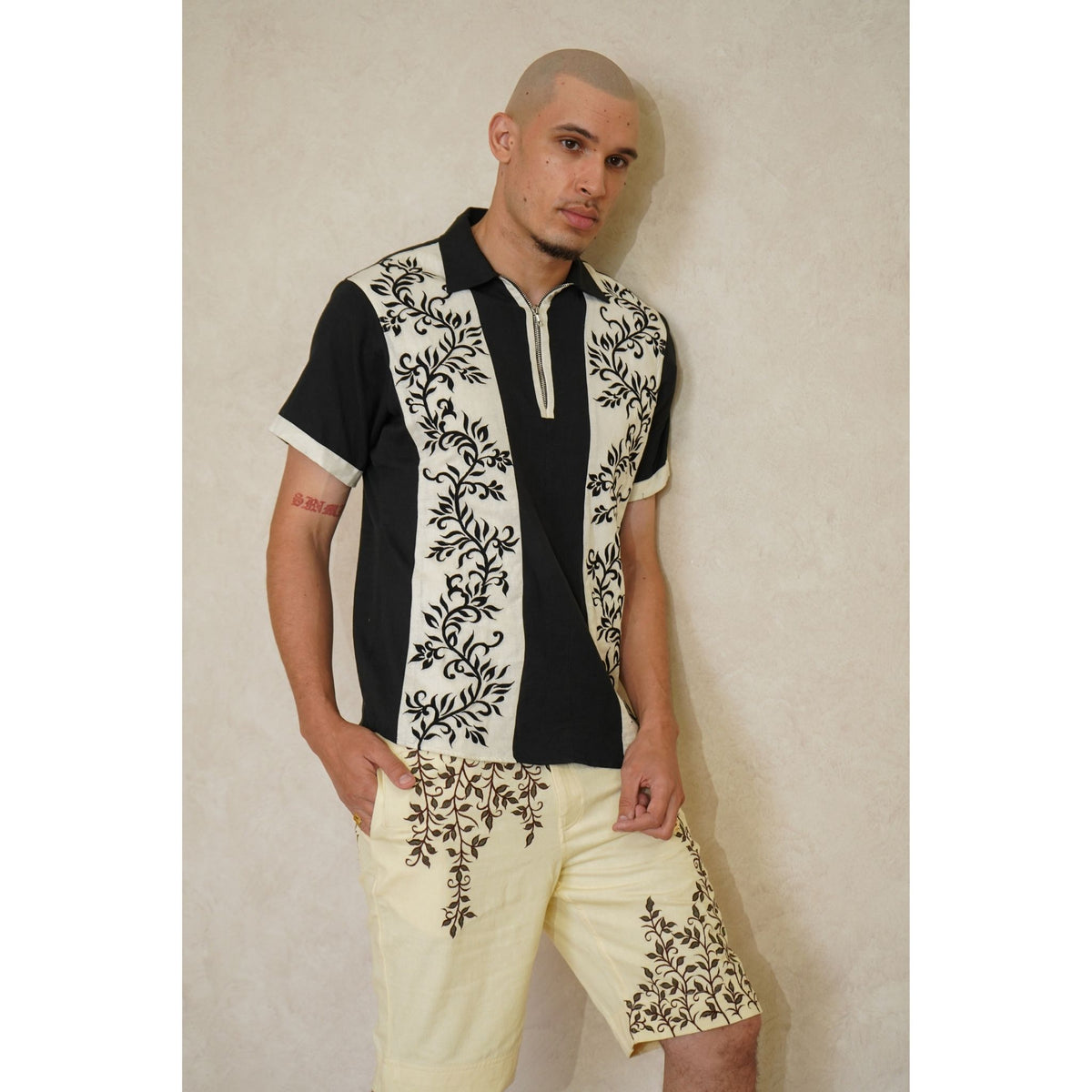 A man with short blond hair showcases INTERNATIONAL APPEAL’s Black Floral-Embroidered Panel Shirt (PR Sample) paired with light floral shorts, standing against a plain, light-colored wall.