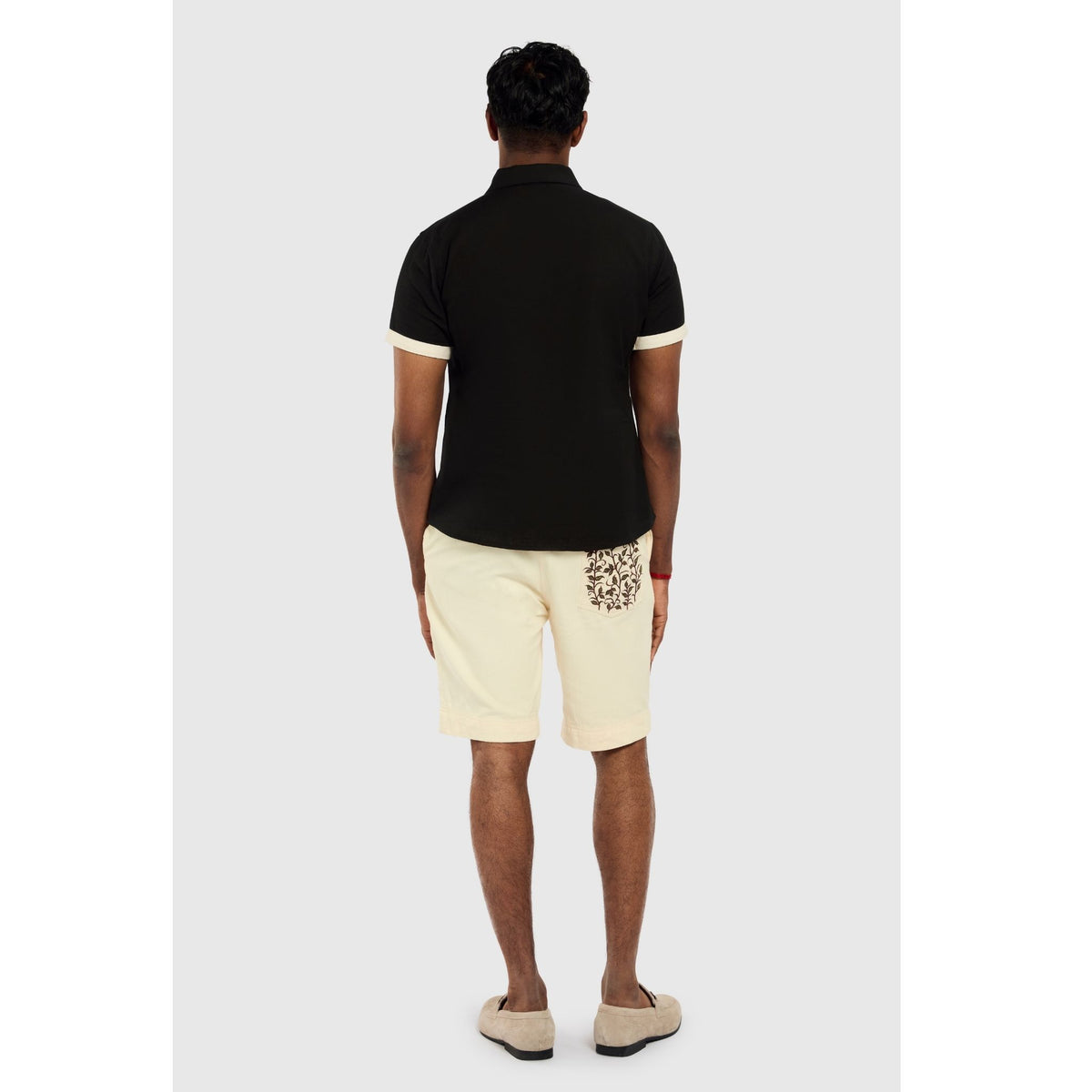 A man stands facing away in a short-sleeve black polo, INTERNATIONAL APPEAL Beige Vine Embroidered Shorts (PR Sample), and beige slip-ons, set against a plain light gray background.