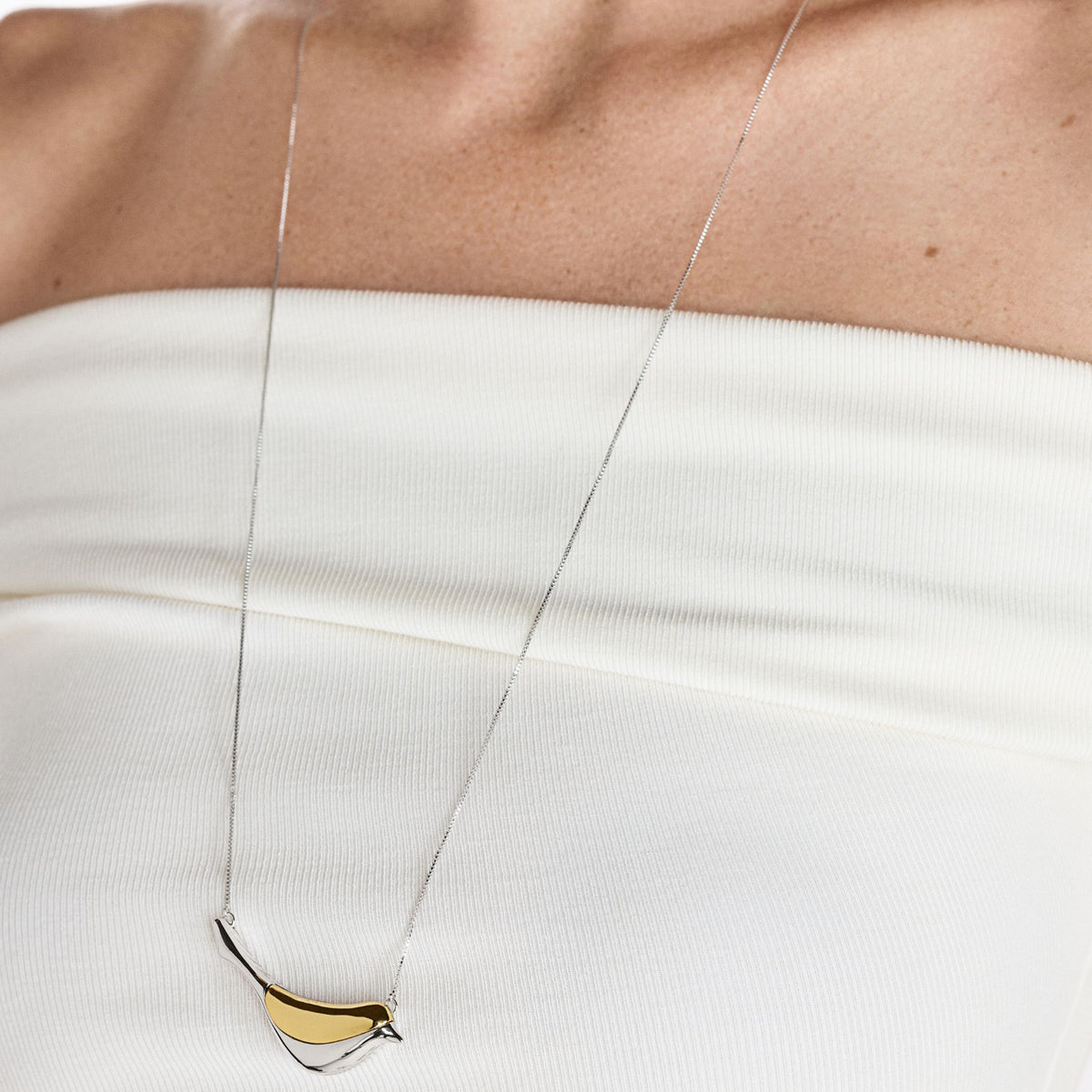 A person wears a strapless white top and the ANNA PRATA Silver Aviva Necklace from the Aviva Collection, featuring a long chain with a curved two-tone silver and gold pendant that resembles a symbolic amulet.