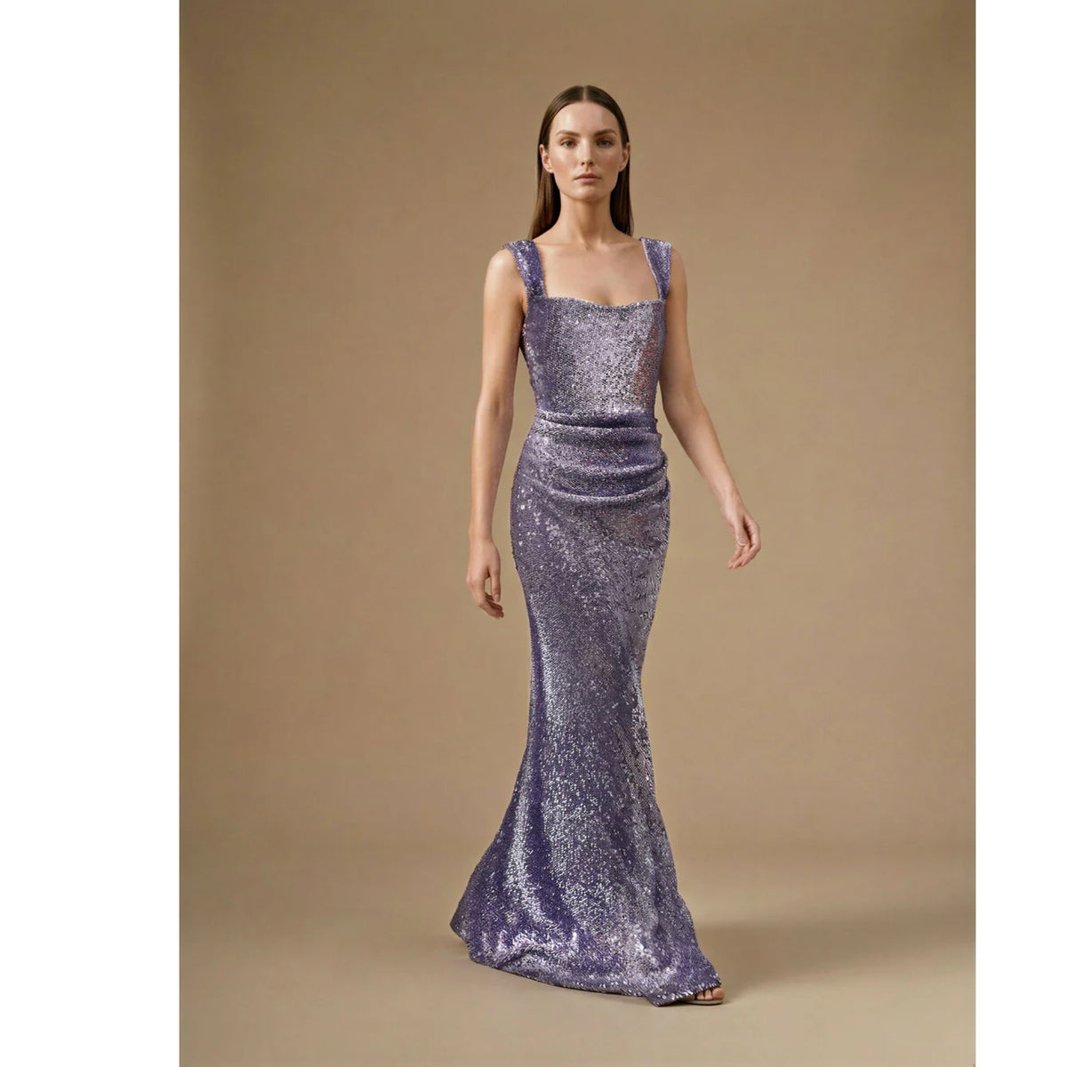 A woman with long, straight brown hair and a neutral expression models the ASHLEY STAMBOULI Doria Dress | PR Sample, a floor-length purple sequin gown with a square neckline and fitted silhouette, against a plain beige background.