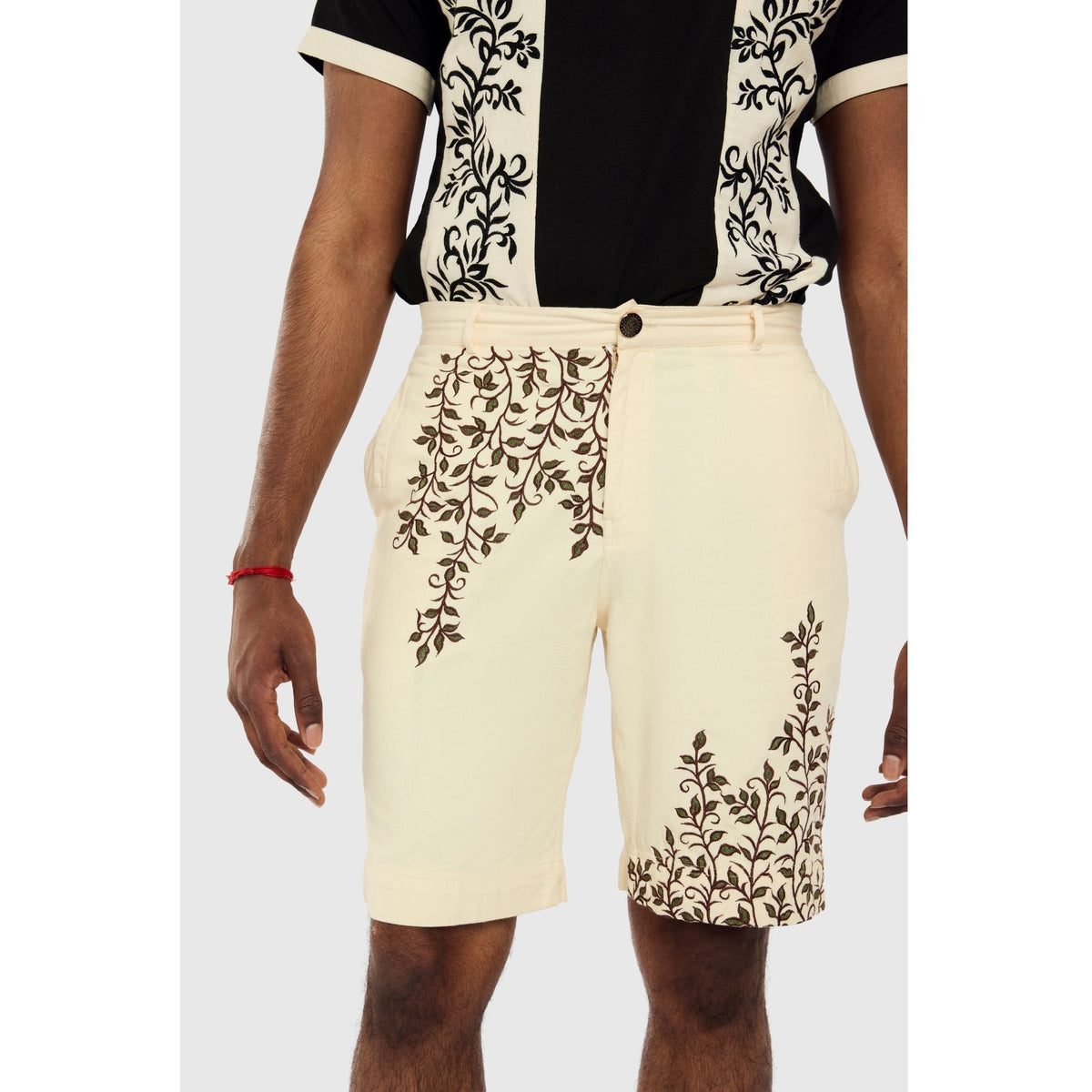 A person models INTERNATIONAL APPEAL's Beige Vine Embroidered Shorts (PR Sample) with a coordinating black and cream shirt, ideal for modern resort wear, standing against a plain light gray background.