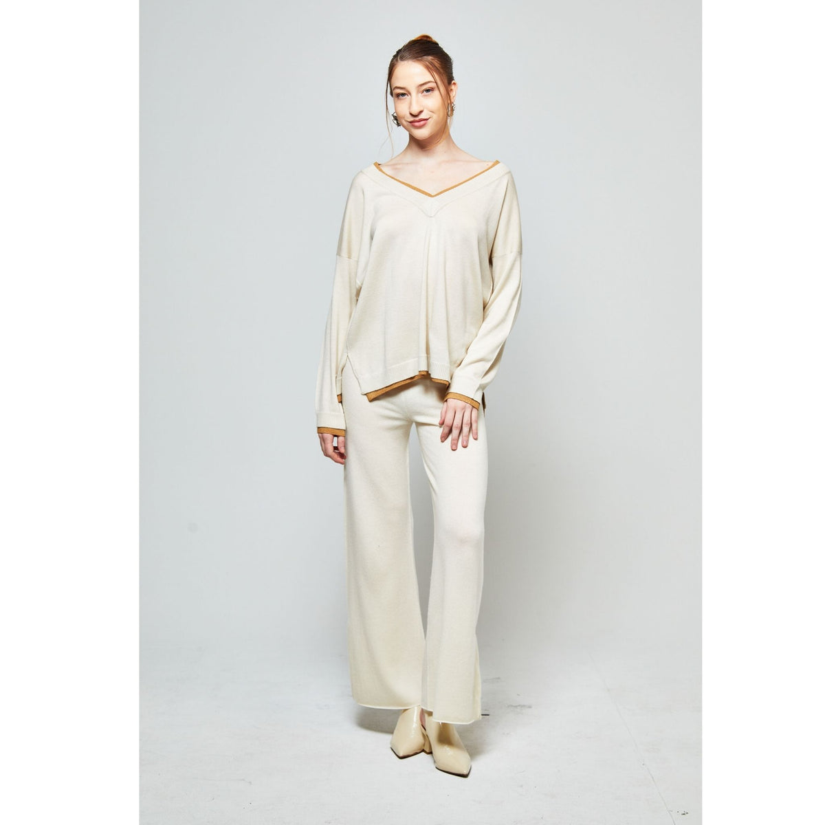 A woman stands against a white background in OO-TO's White with Beige Lapis Oversized V-Neck Sweater, paired with matching wide-leg pants and cream shoes. Her hair is tied back. The cozy sweater is handcrafted in Nepal.
