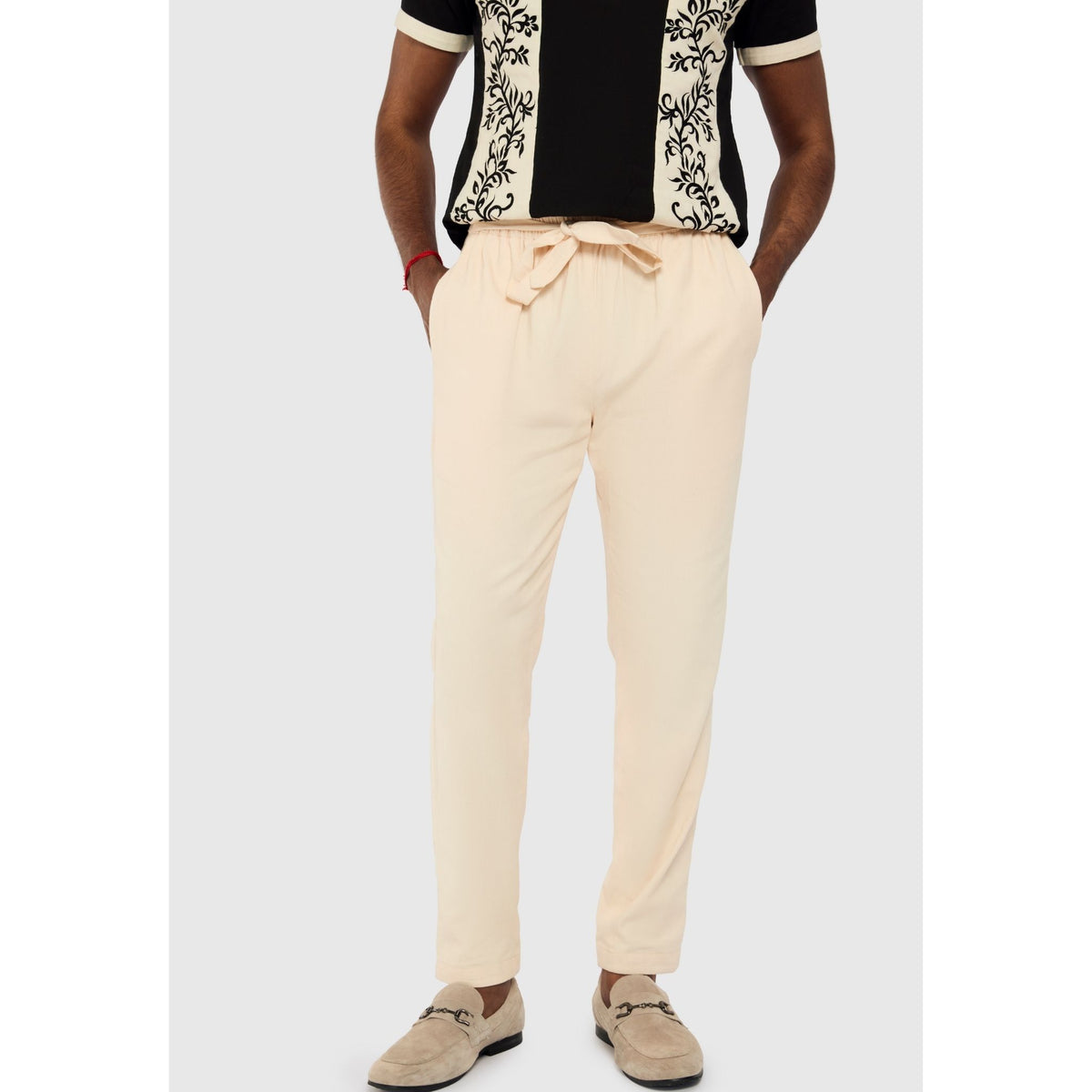 A man models INTERNATIONAL APPEAL’s Beige Linen Tailored Pants | PR Sample, styled with tan loafers and a black-and-white patterned short-sleeve shirt, epitomizing modern resort tailoring against a light gray backdrop.