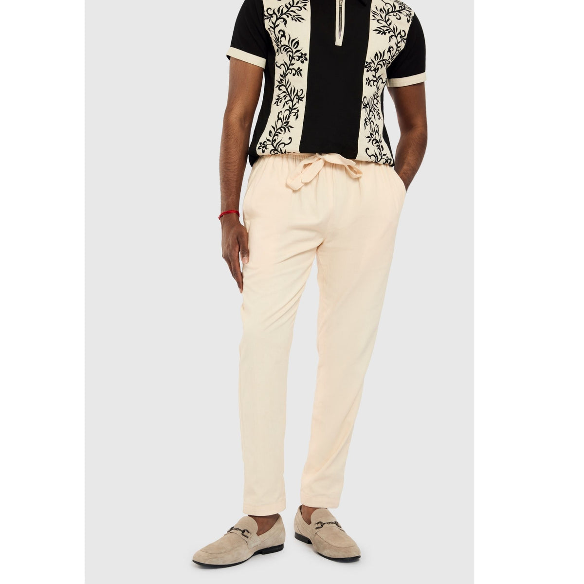 A person in INTERNATIONAL APPEAL's Beige Linen Tailored Pants (PR Sample), a black and beige patterned short-sleeve shirt, and beige loafers with black detailing stands with hands in pockets against a plain background.