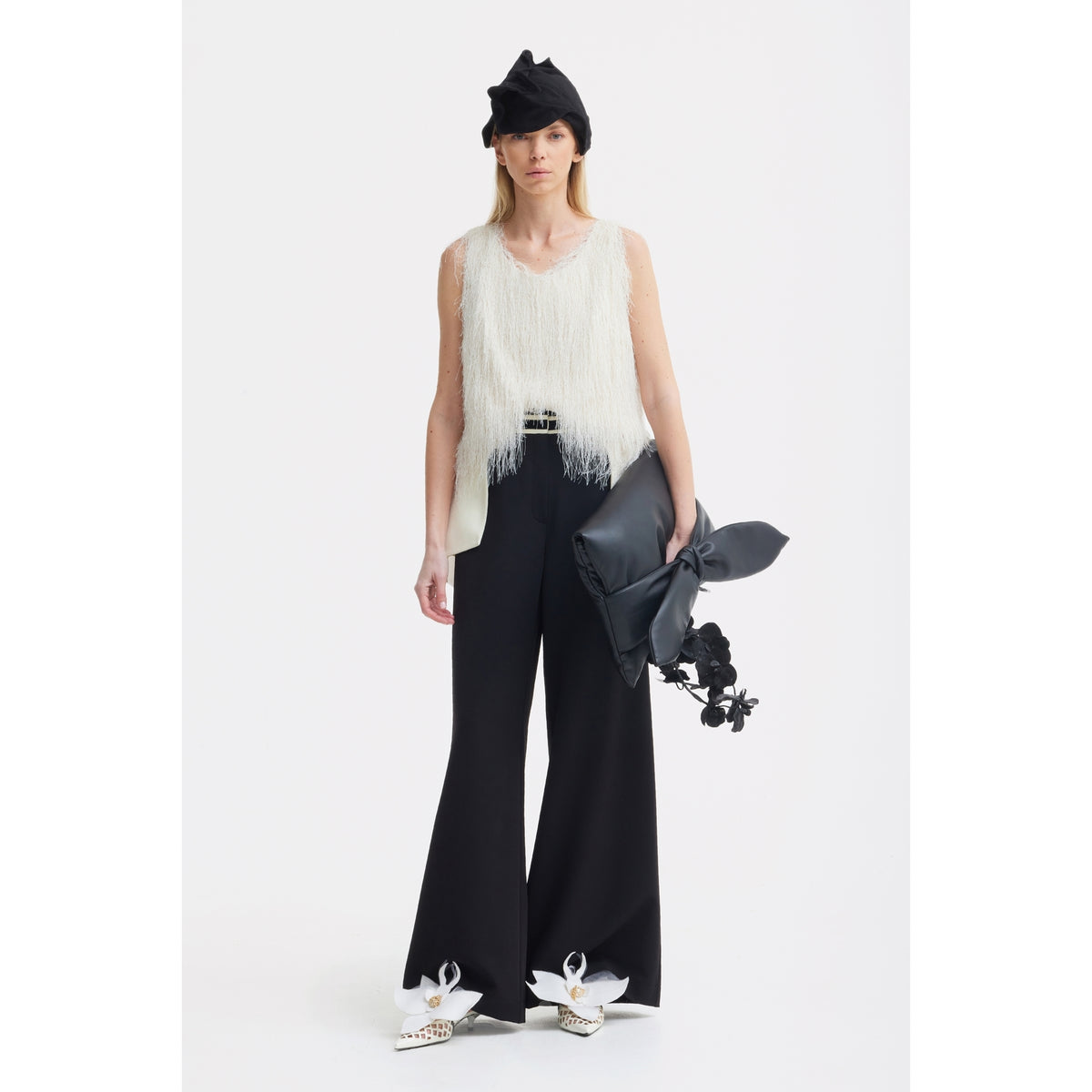 A woman stands against a white background wearing JULIA ALLERT's Statement Waist Wide-Leg Trousers in black, a white fringed sleeveless top, white bow-accented shoes, a black hat, and holding a floral-accented black clutch.