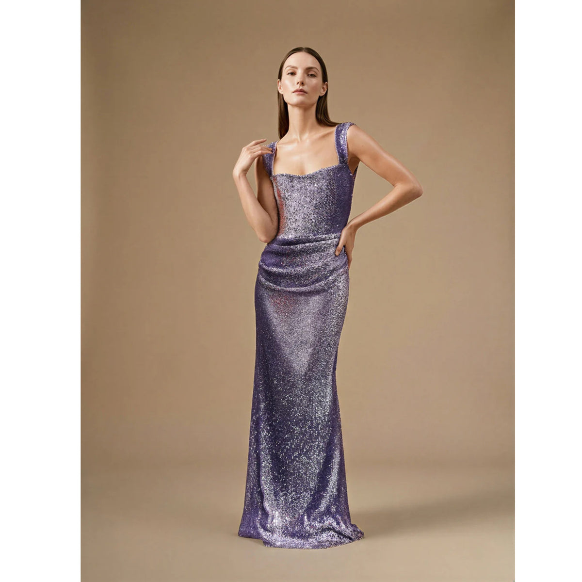 A woman stands confidently in the floor-length, sleeveless Doria Dress by ASHLEY STAMBOULI, a fitted mermaid gown with wide straps and shimmering lavender fabric that catches the light, set against a plain beige background.
