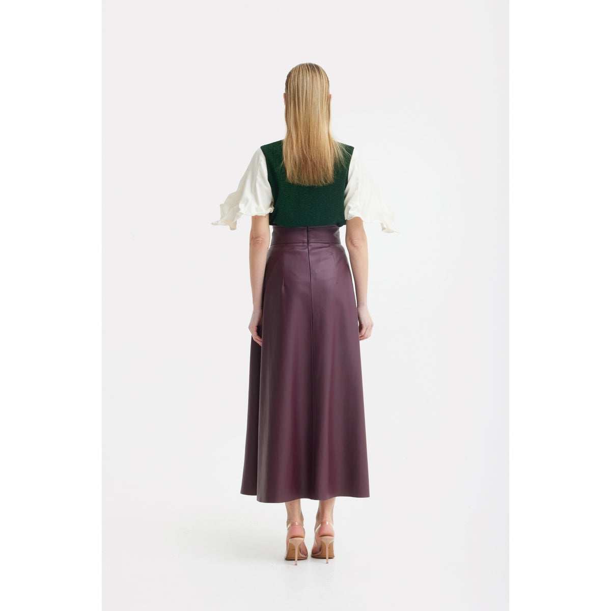 A woman with straight blonde hair stands against a white background, wearing the JULIA ALLERT Green Lurex Knit Top With Adjustable Satin Sleeves and a high-waisted, ankle-length plum skirt, paired with nude high-heeled shoes.