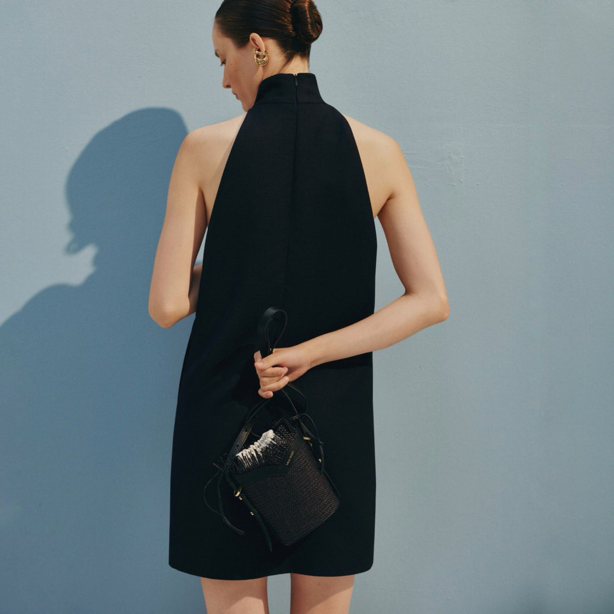 Woman in a black dress holding a black handbag against a light blue wall