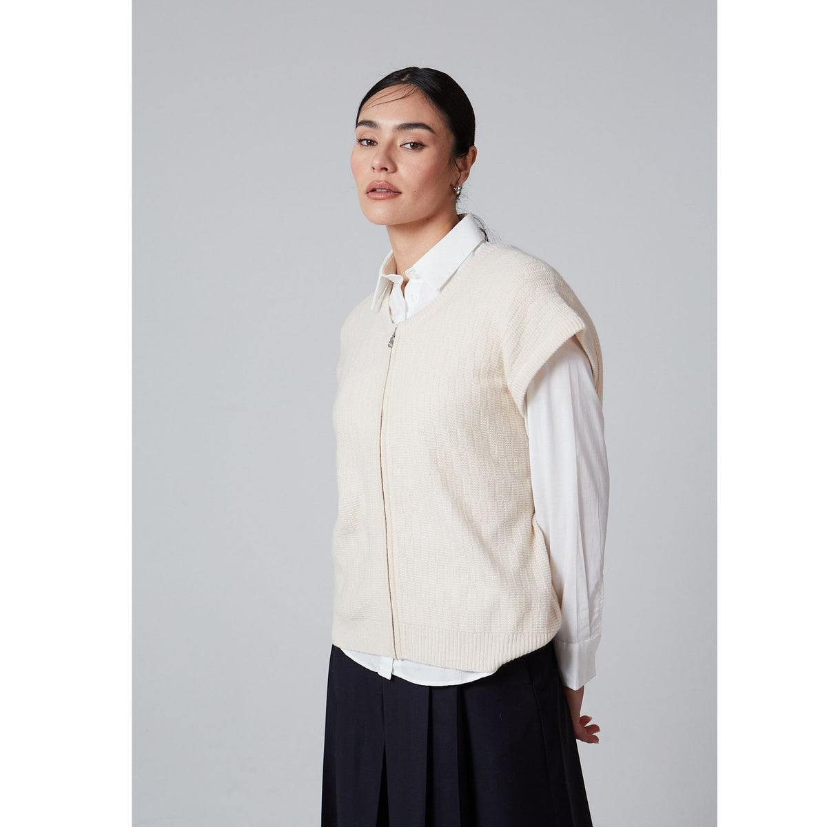 A woman with dark hair tied back wears the OO-TO White Unisex Sleeveless Vest Arnau over a white shirt and black skirt, standing against a light gray background—showcasing elegant transitional layering.