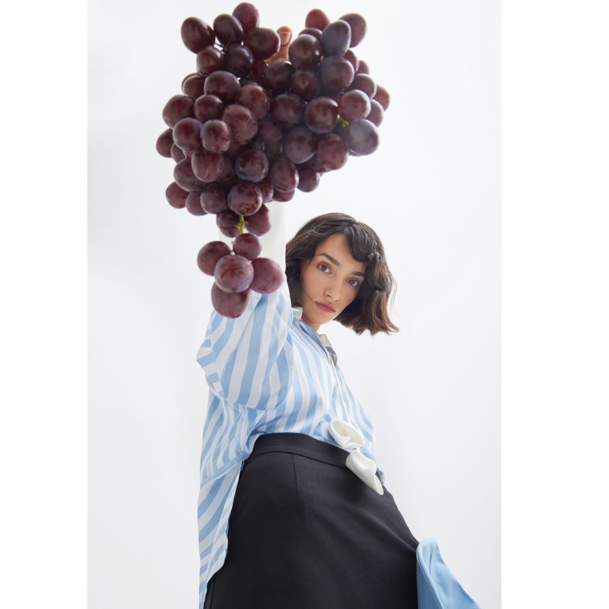 A woman models the Smart Elegance – Silk-Touch Striped Long-Sleeve Blouse in Light Blue by JULIA ALLERT, holding red grapes against a white background.