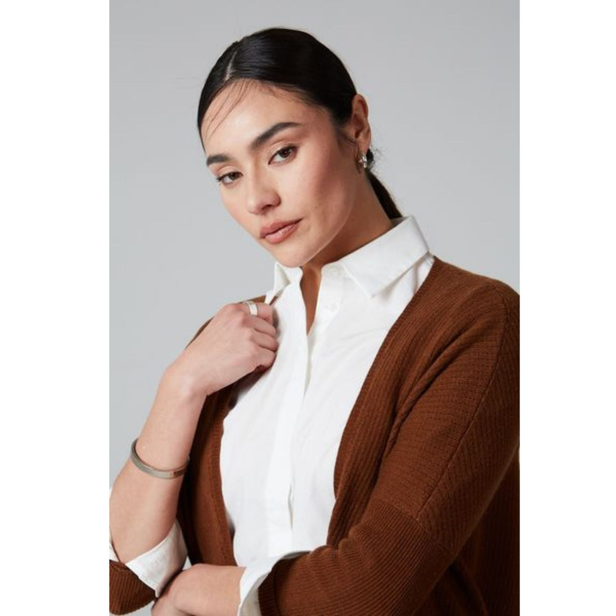 A woman with dark hair tied back wears the OO-TO Syrup Brown Lillien Open Cardigan over a white collared shirt. She confidently faces the camera, touching her collar, against a plain light gray background.
