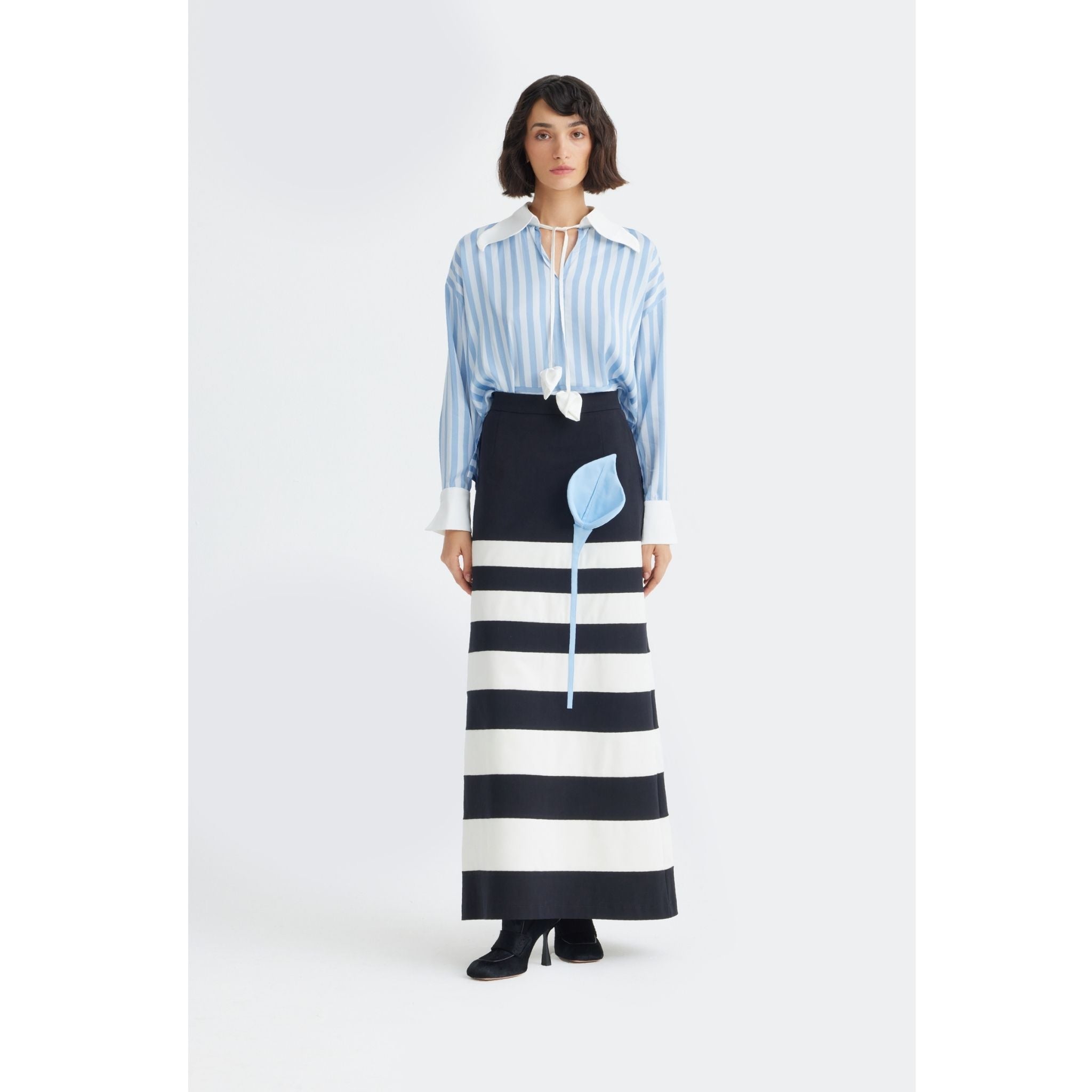 A woman stands against a plain background wearing the JULIA ALLERT Smart Elegance Silk-Touch Striped Long-Sleeve Blouse in light blue, paired with a long black skirt with wide white stripes, black boots, and a light blue leaf accessory.