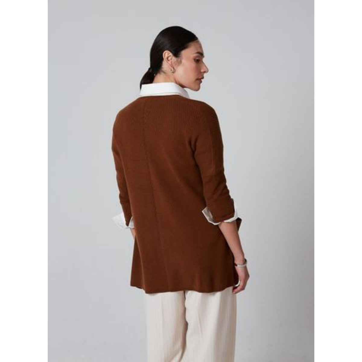A woman with dark hair in a low bun stands facing away, wearing the OO-TO Syrup Brown Lillien Open Cardigan over a white collared shirt and cream trousers. This cardigan is handcrafted in Nepal for a look of timeless elegance.