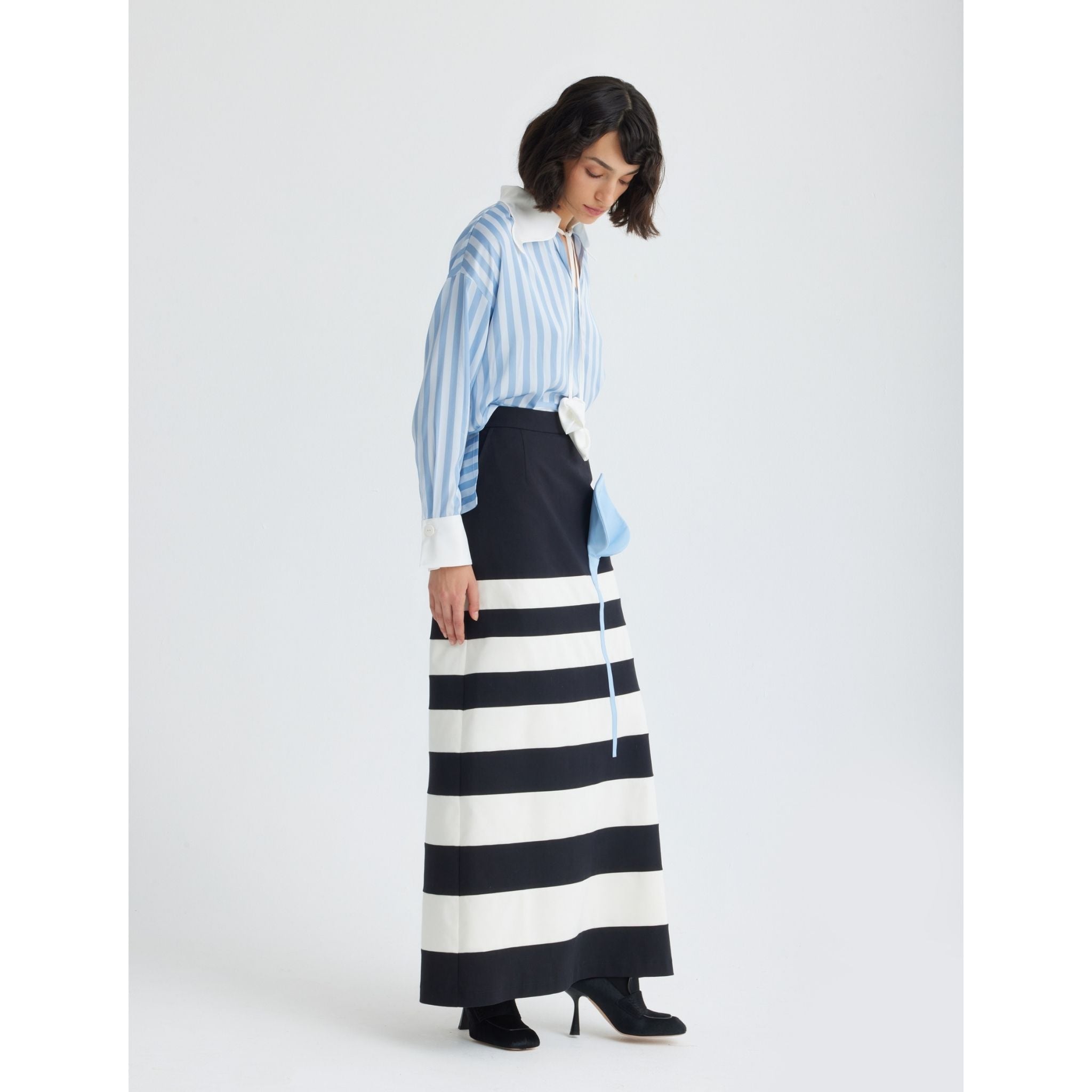 A woman stands against a white background, wearing the JULIA ALLERT Smart Elegance Silk-Touch Striped Long-Sleeve Blouse in light blue, paired with a striped skirt, bow belt, and black ankle boots. She looks down, her hair in loose waves.